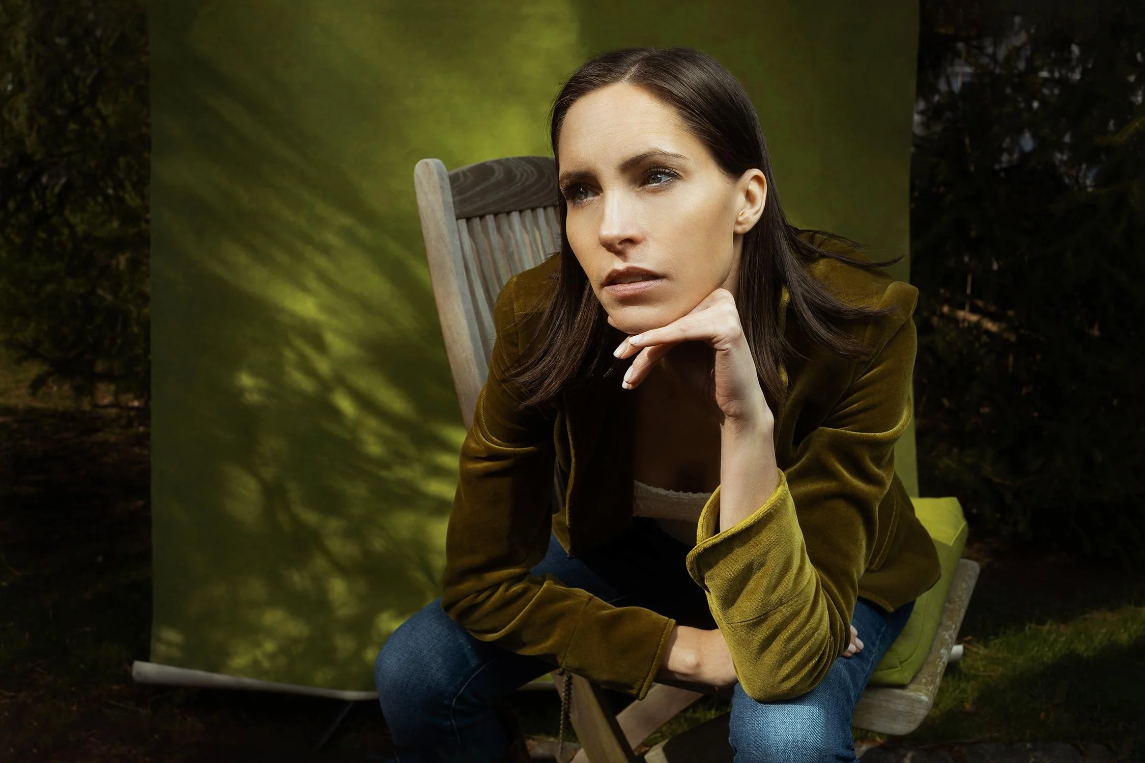 A woman with dark hair in a brown jacket sitting outdoors on a wooden chair with a green cushion, resting her chin on her hand, looking thoughtful, with a background of foliage and a green backdrop.