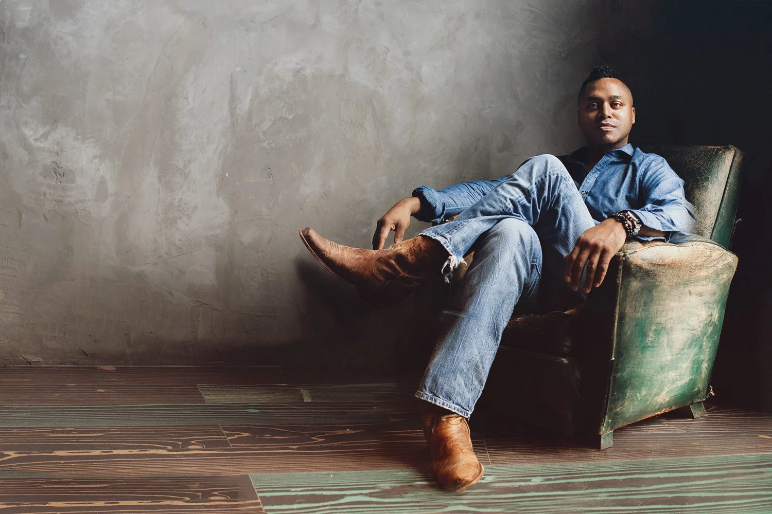 A man sitting in a vintage armchair with his legs crossed, wearing a blue denim shirt, jeans, and brown boots, with bracelets on his left wrist, against a gray textured wall and wooden floor.