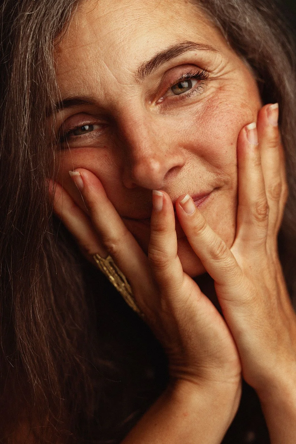 A close-up of a woman with dark hair, touching her face with her fingers, showing her expression and facial features.