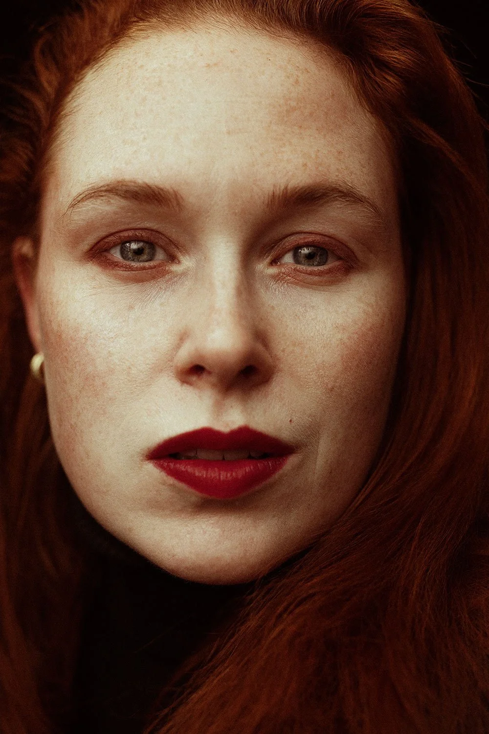 Close-up of a woman with red hair and blue eyes, wearing red lipstick and a small earring.