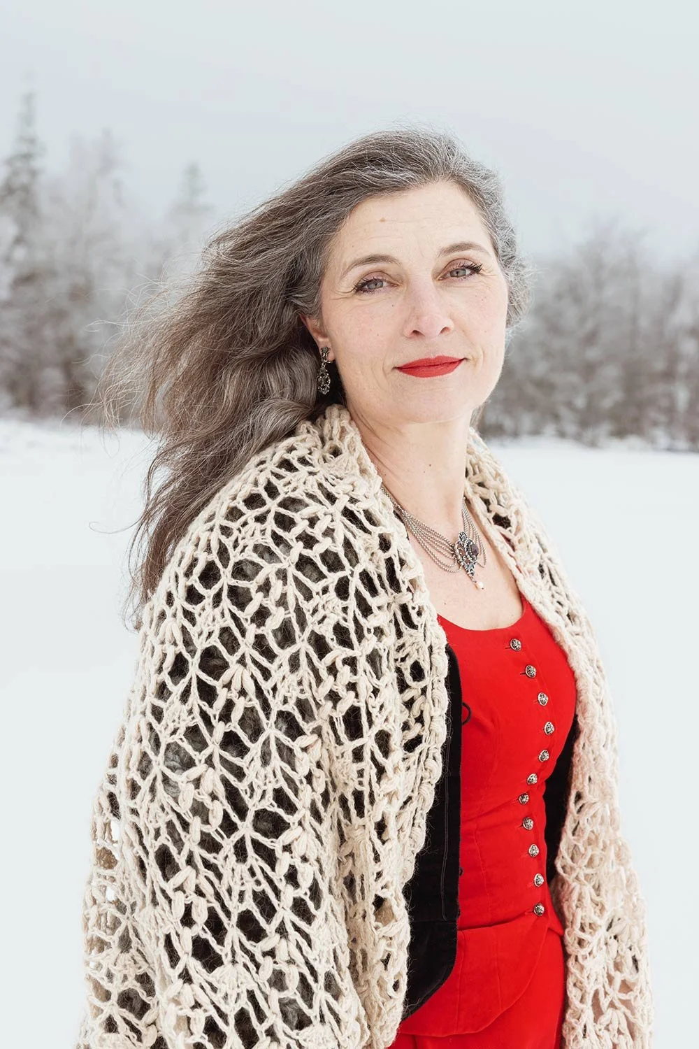 A woman with gray hair and red lipstick standing outdoors in a snowy landscape, wearing a beige crocheted shawl, a red blouse with buttons, and jewelry.