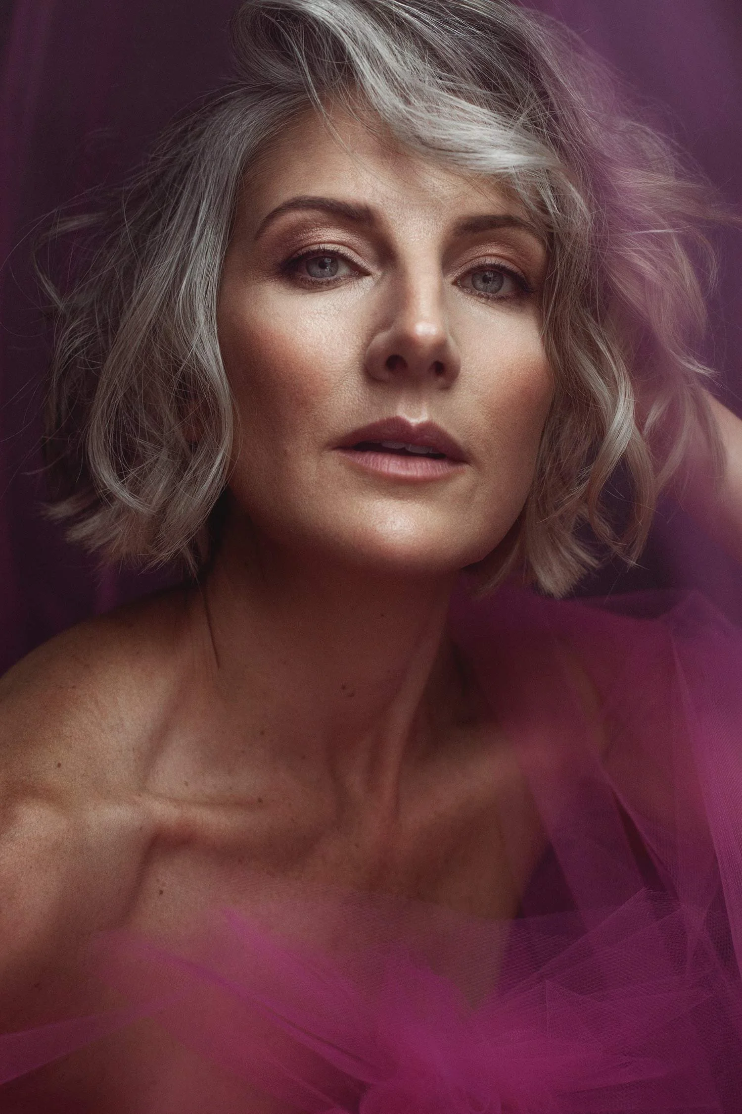 A woman with short, wavy silver hair, blue eyes, and light makeup, looking directly at the camera against a dark background, with pink sheer fabric in the foreground.