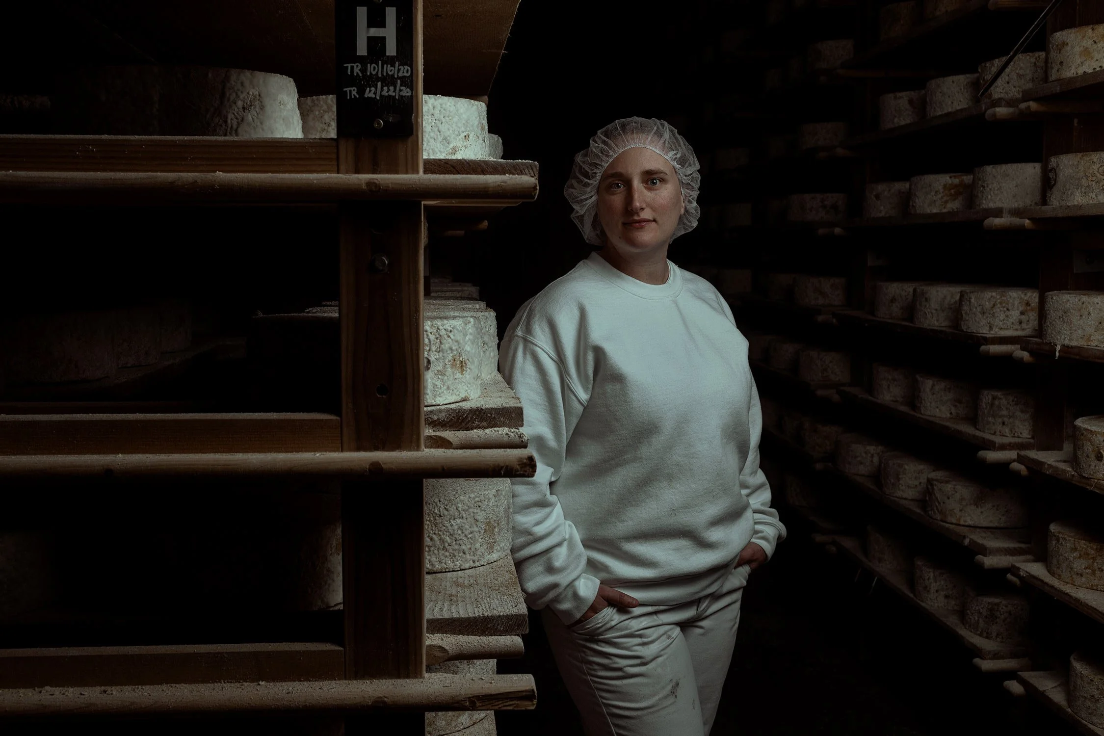 A woman wearing a hairnet and white clothing stands in a cheese aging cellar surrounded by rounds of aging cheese on wooden shelves.