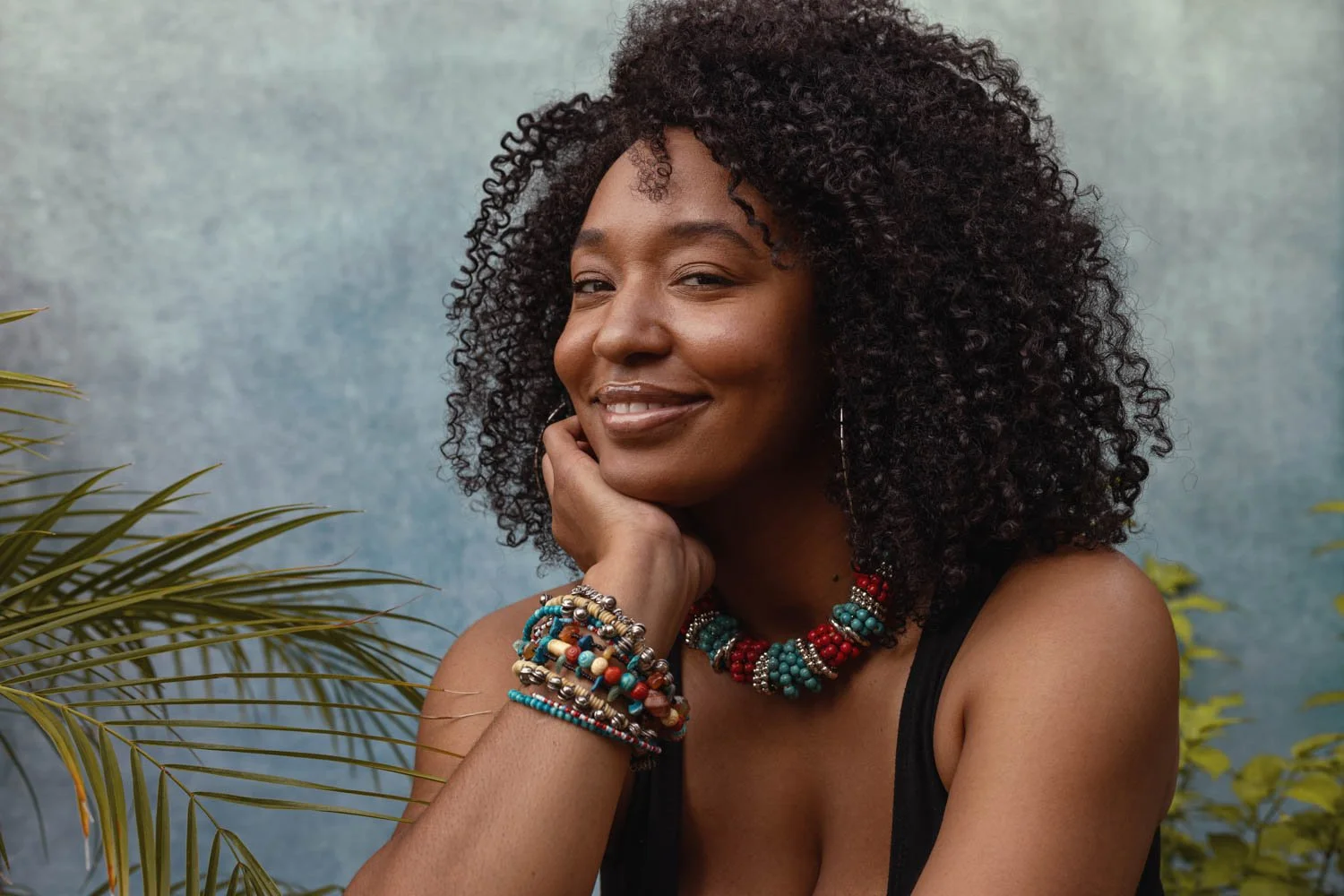 A woman with natural curly hair smiles softly, resting her chin on her hand, wearing colorful beaded jewelry including a necklace and bracelets, with plants and a light blue textured background.