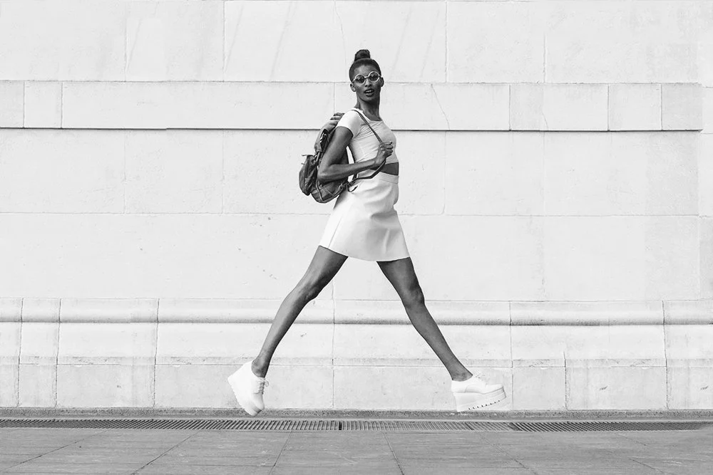 A young woman wearing sunglasses, a small top, a skirt, and platform shoes, carrying a backpack, walking briskly in front of a plain wall.
