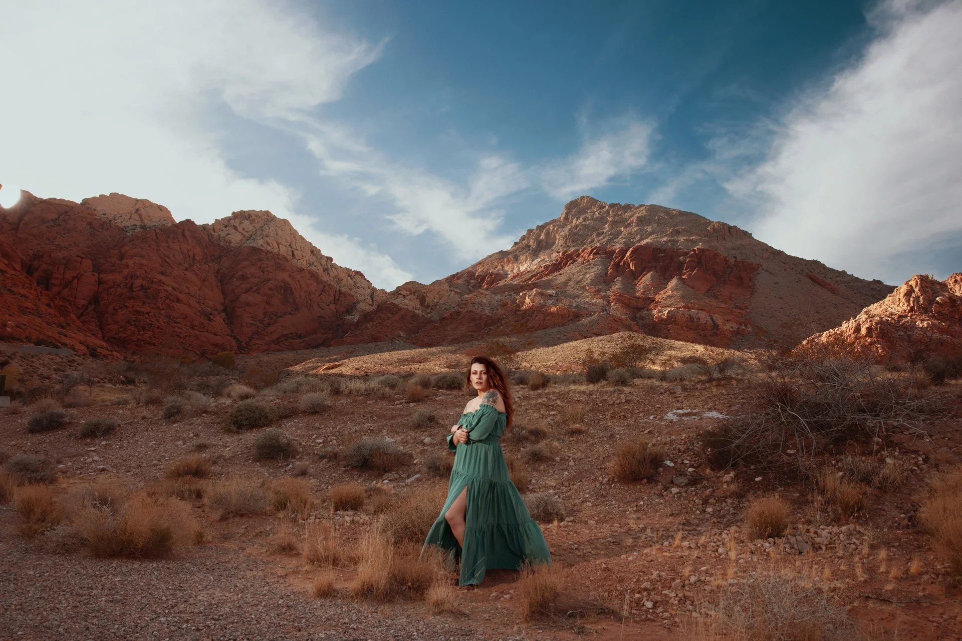 A woman in a green dress stands in a desert landscape with red mountains and a blue sky with clouds in the background.