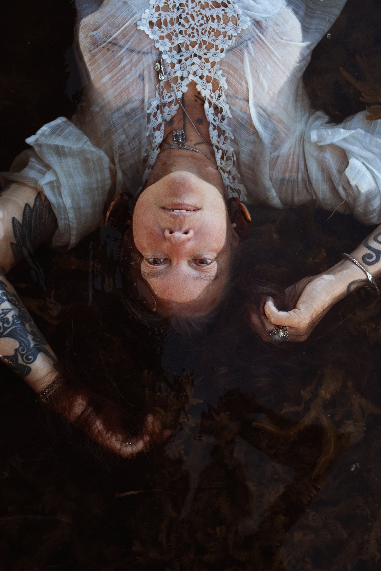 A woman with tattoos and jewelry lying in water, wearing a white lace top, looking up at the camera.