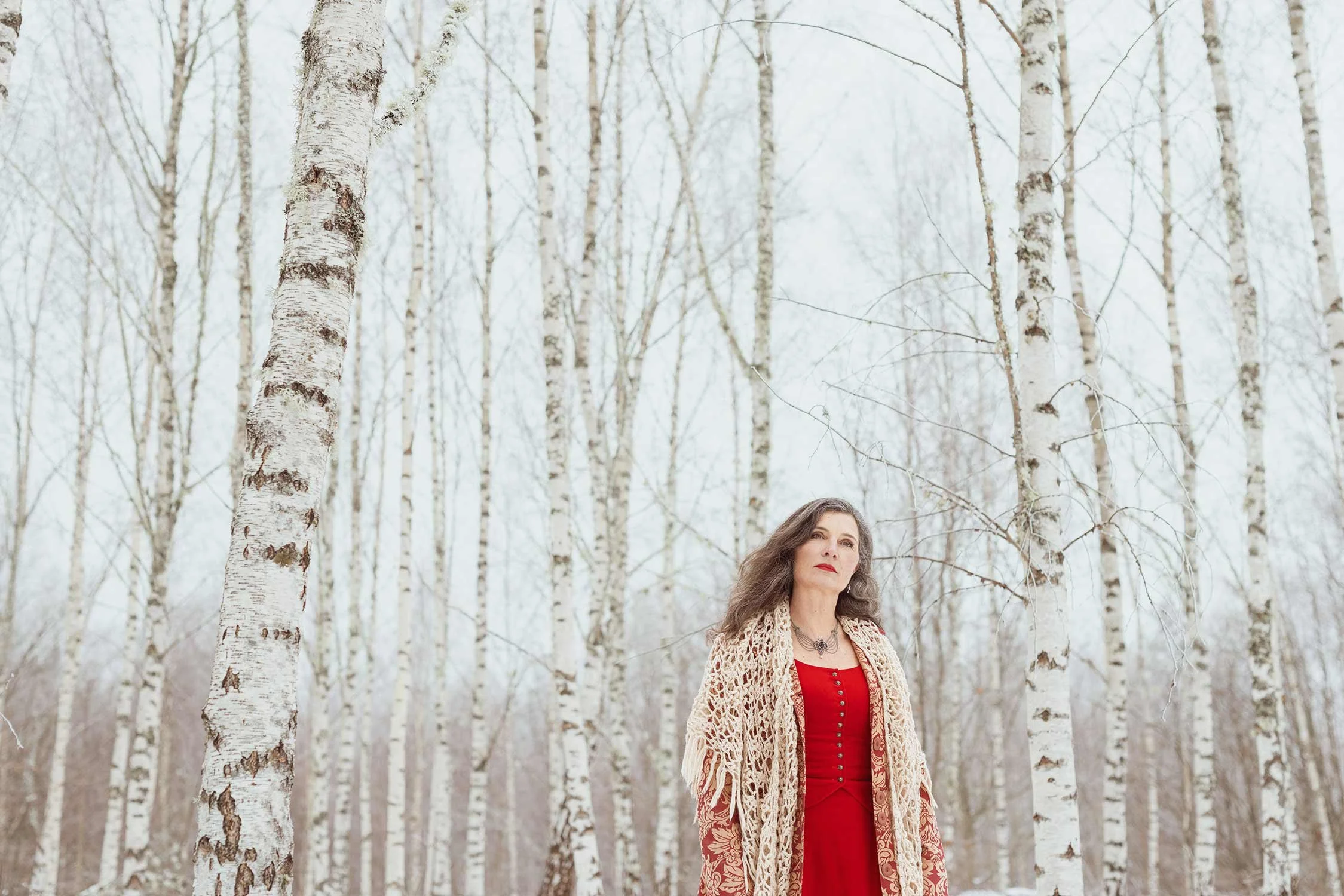 A woman with long gray hair dressed in a red dress and a beige crochet shawl stands in a snow-covered forest of white birch trees.