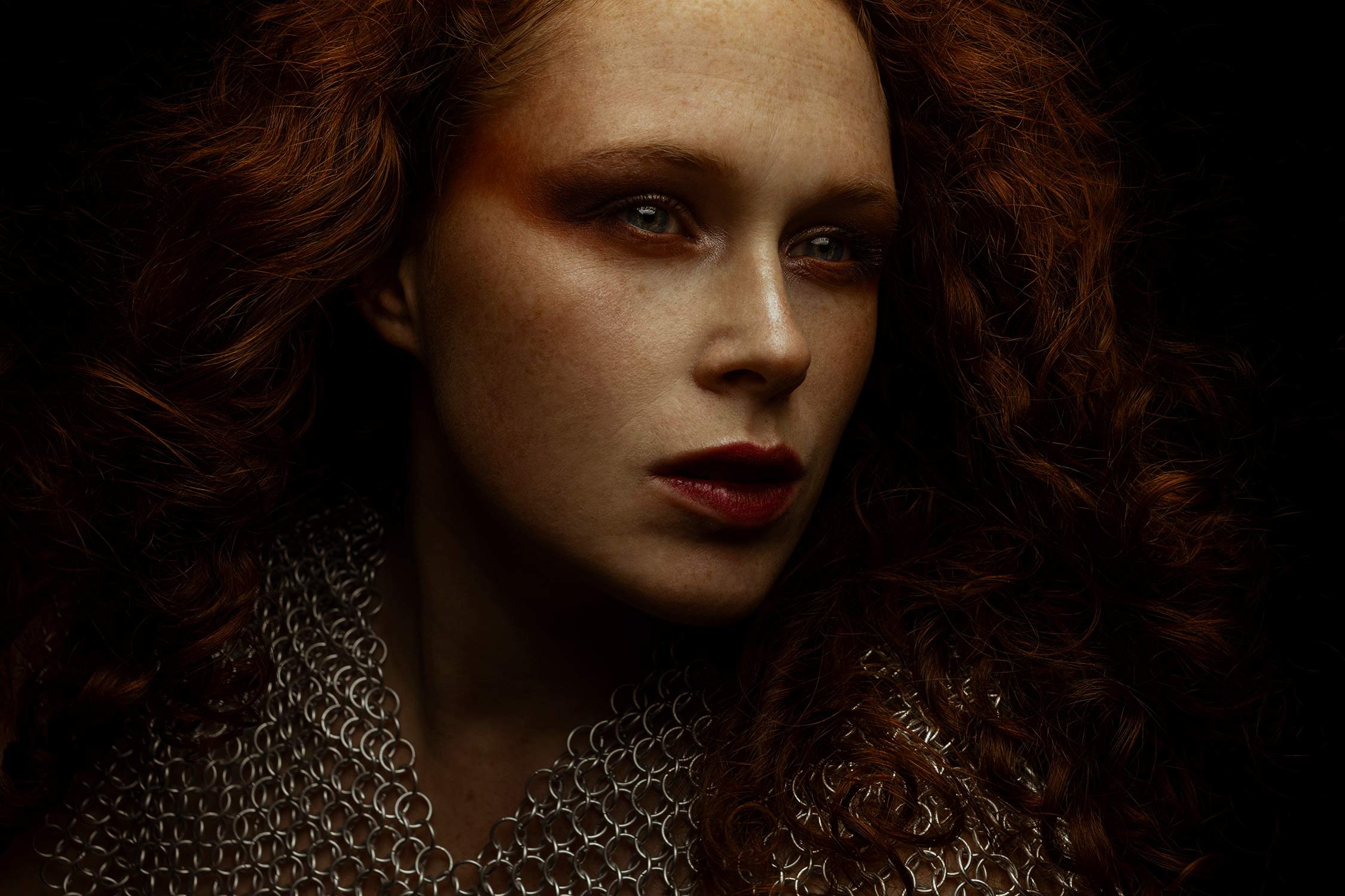 Close-up portrait of a woman with red curly hair and fair complexion, wearing a chainmail necklace, against a dark background.