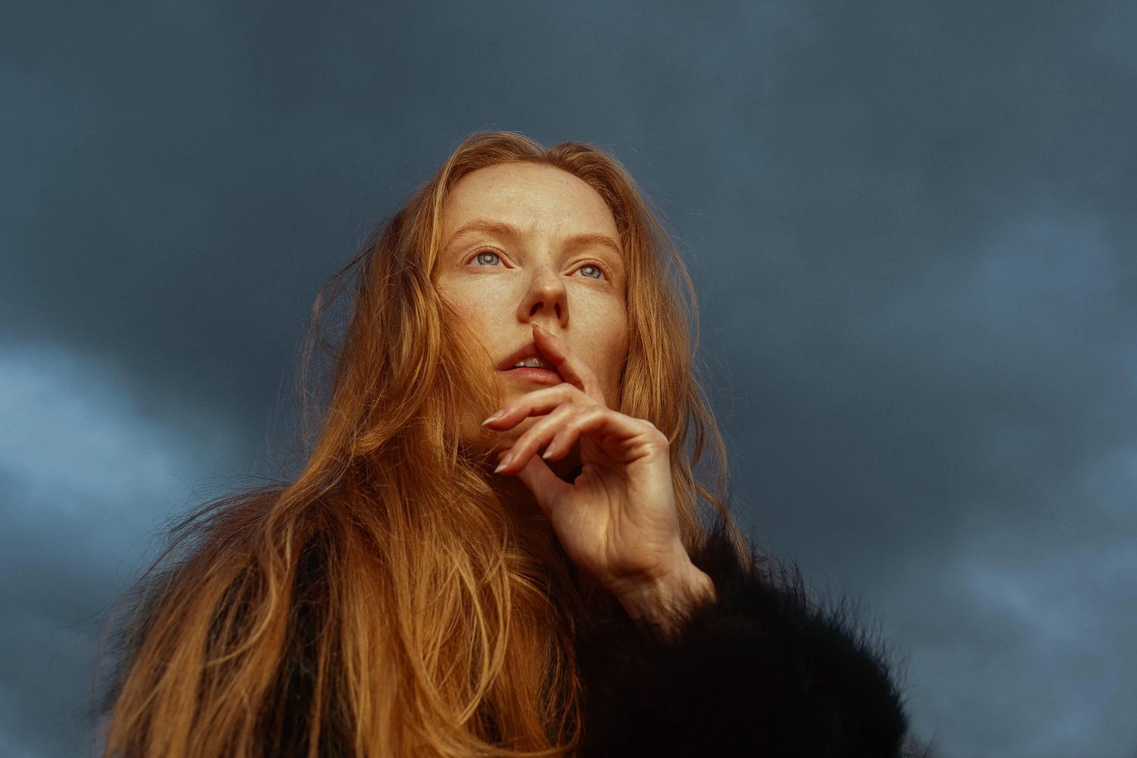 A woman with long red hair and fair skin holding her finger to her lips, looking upwards against a dark, cloudy sky background.