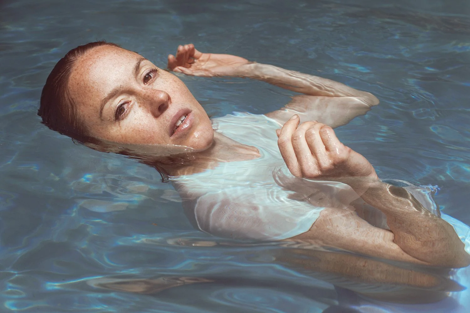 A woman with wet red hair and freckles floats on her back in a pool, looking at the camera with a relaxed expression.