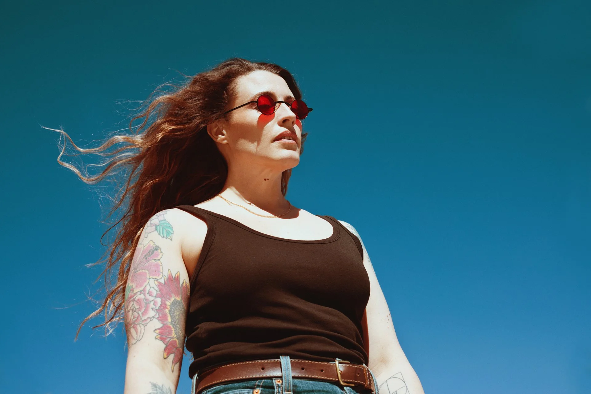 Woman with long hair wearing red sunglasses, a black tank top, and has tattoos on her arm, standing outdoors against a clear blue sky.