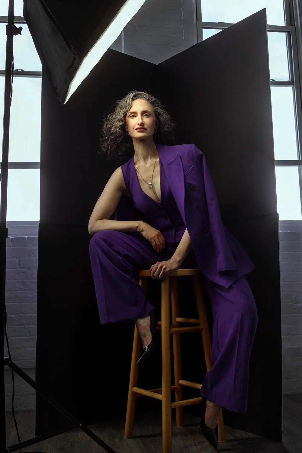 A woman in a purple suit sitting on a wooden stool in front of a black backdrop, with large windows behind her, in a studio setting.