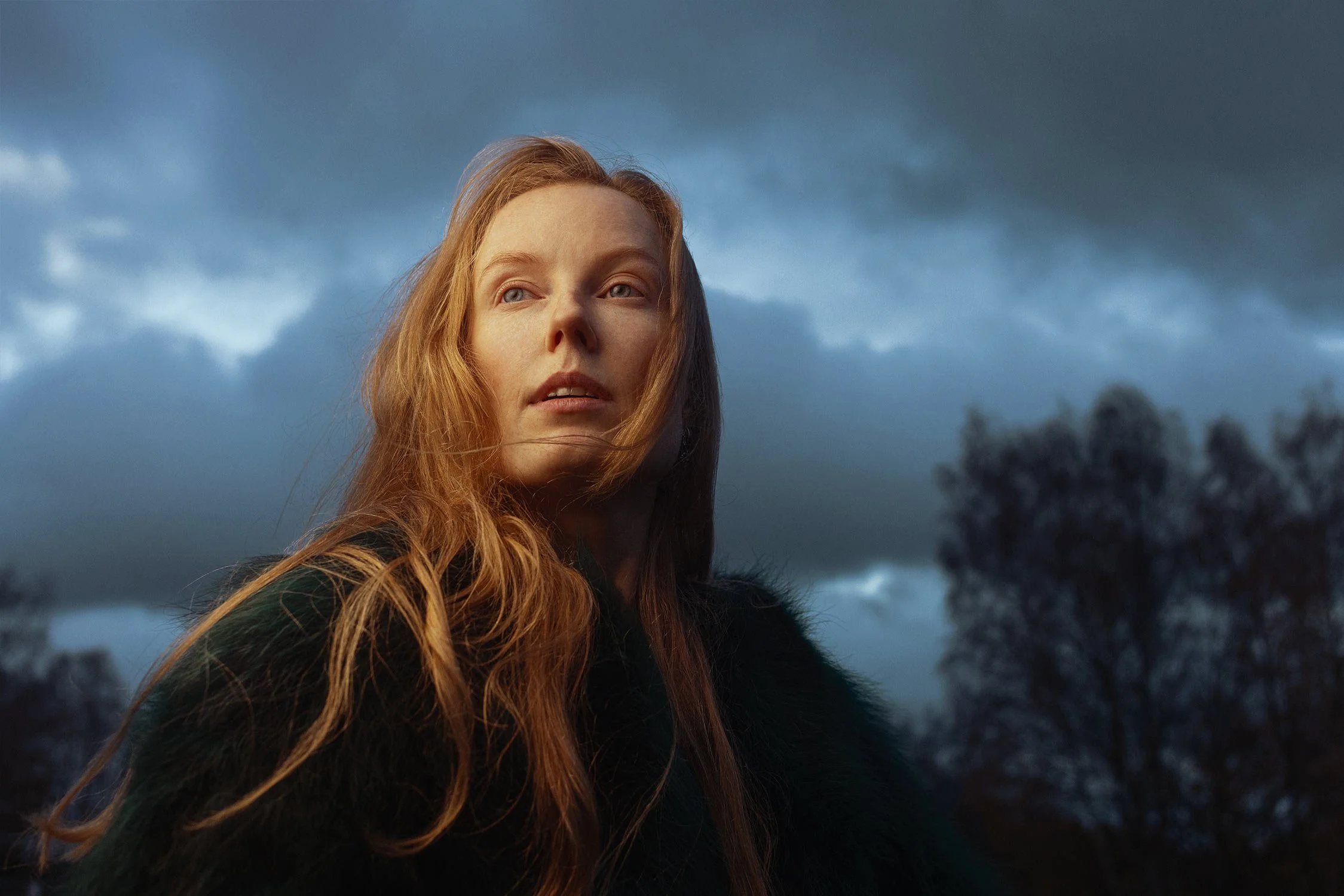 A woman with long red hair and a dark coat looks into the distance against a cloudy sky, with trees in the background.