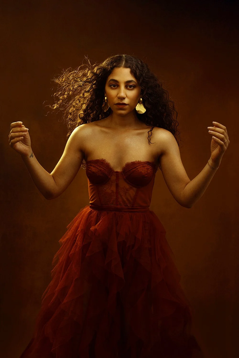 A woman with curly hair wearing a strapless red dress with a tulle skirt, standing against a dark brown background.