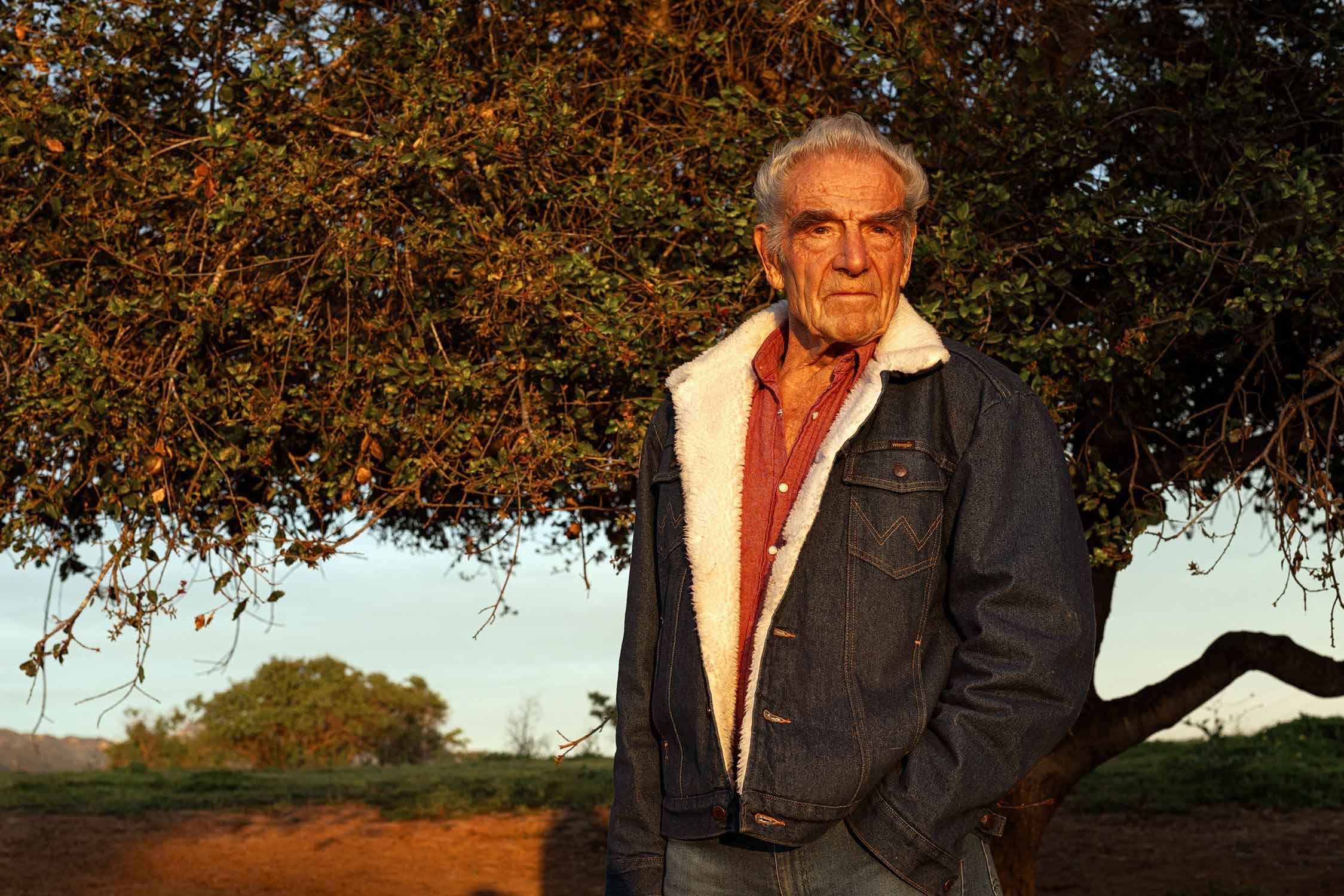 An elderly man stands outdoors near a tree during sunset, wearing a denim jacket with a shearling lining and a red shirt.