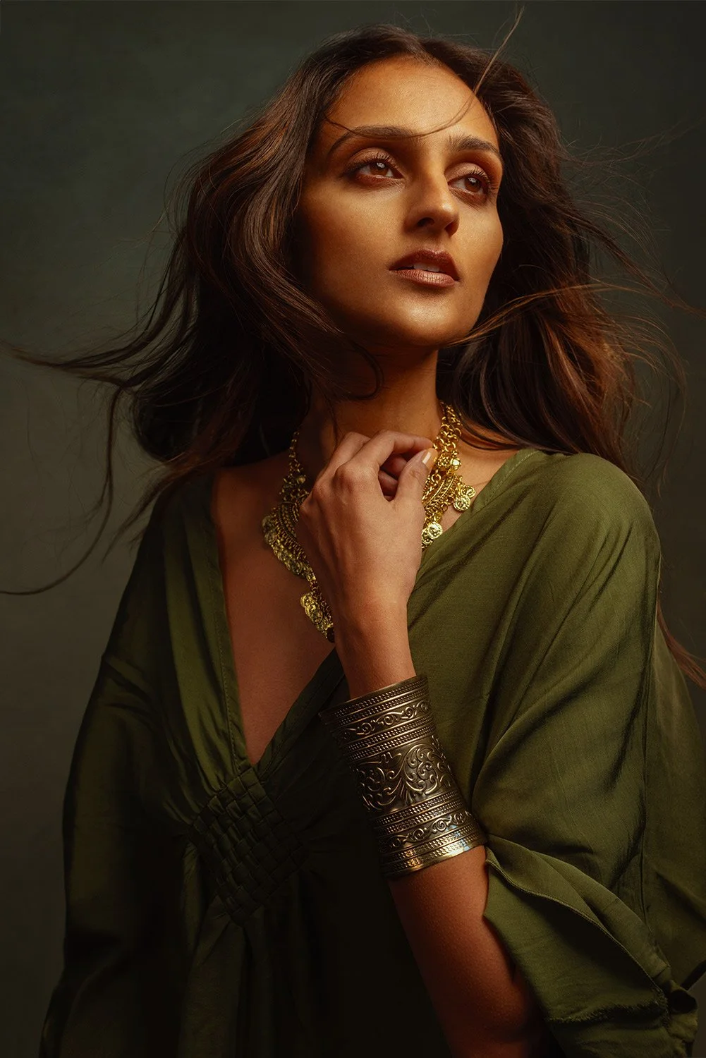 A woman with wavy brown hair wearing a green dress and gold jewelry, including a necklace and bangles, posing with her hand near her neck against a dark background.