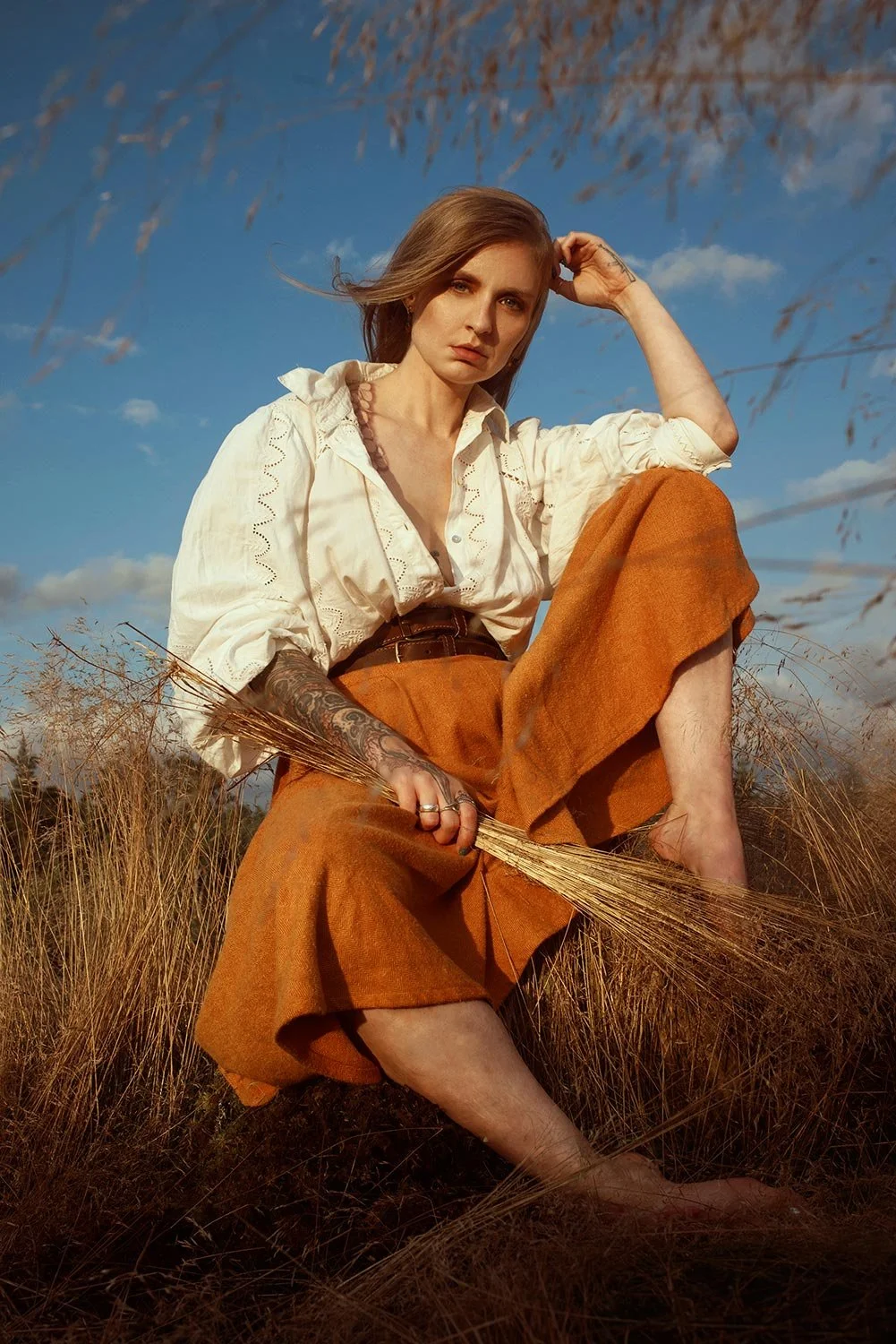 A woman sitting in a field of dry grass during sunset, wearing an off-white blouse with embroidery and brown high-waisted pants, holding a dried grass stalk, with a cloudy sky in the background.