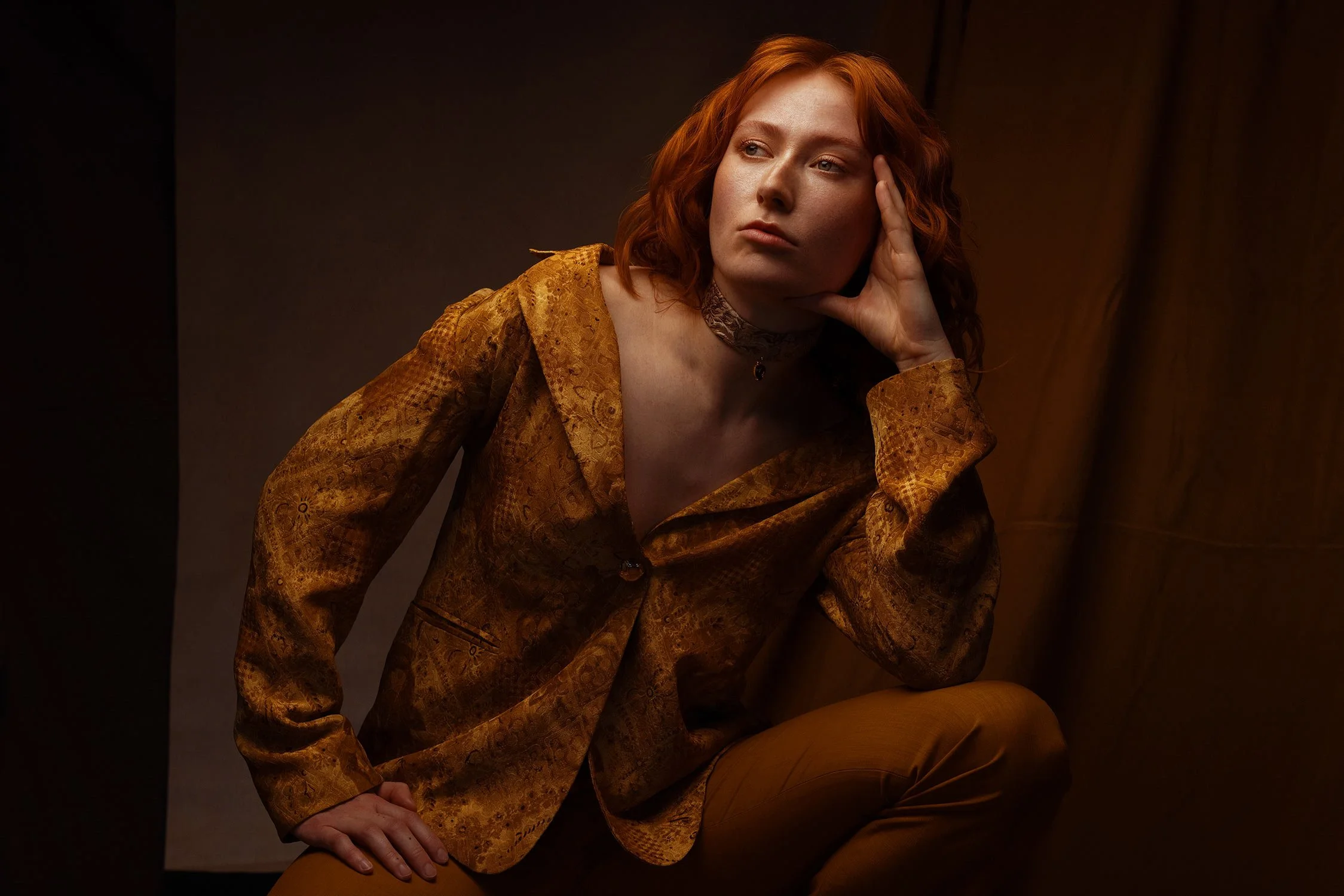 A woman with red hair wearing a patterned brown blazer and pants, posed with her head resting on her hand against a dark background.