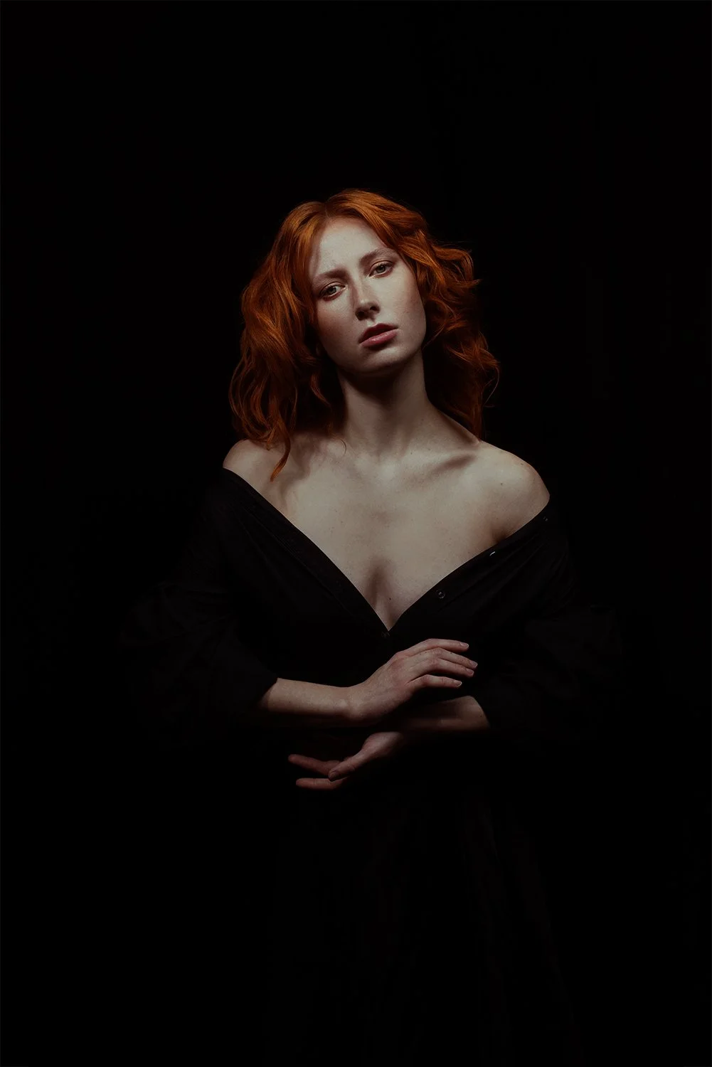 Portrait of a red-haired woman with fair skin wearing a black off-the-shoulder top against a black background.