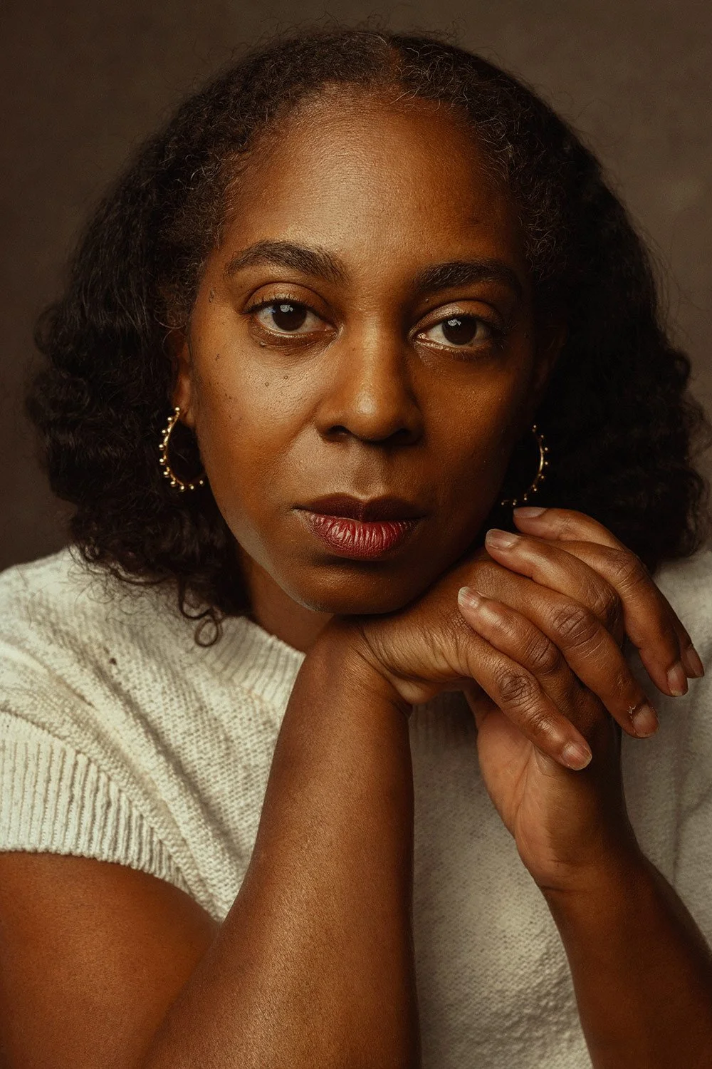 Close-up portrait of a black woman with short, curly black hair, wearing gold hoop earrings and a beige knit top, resting her chin on her hands against a dark background.