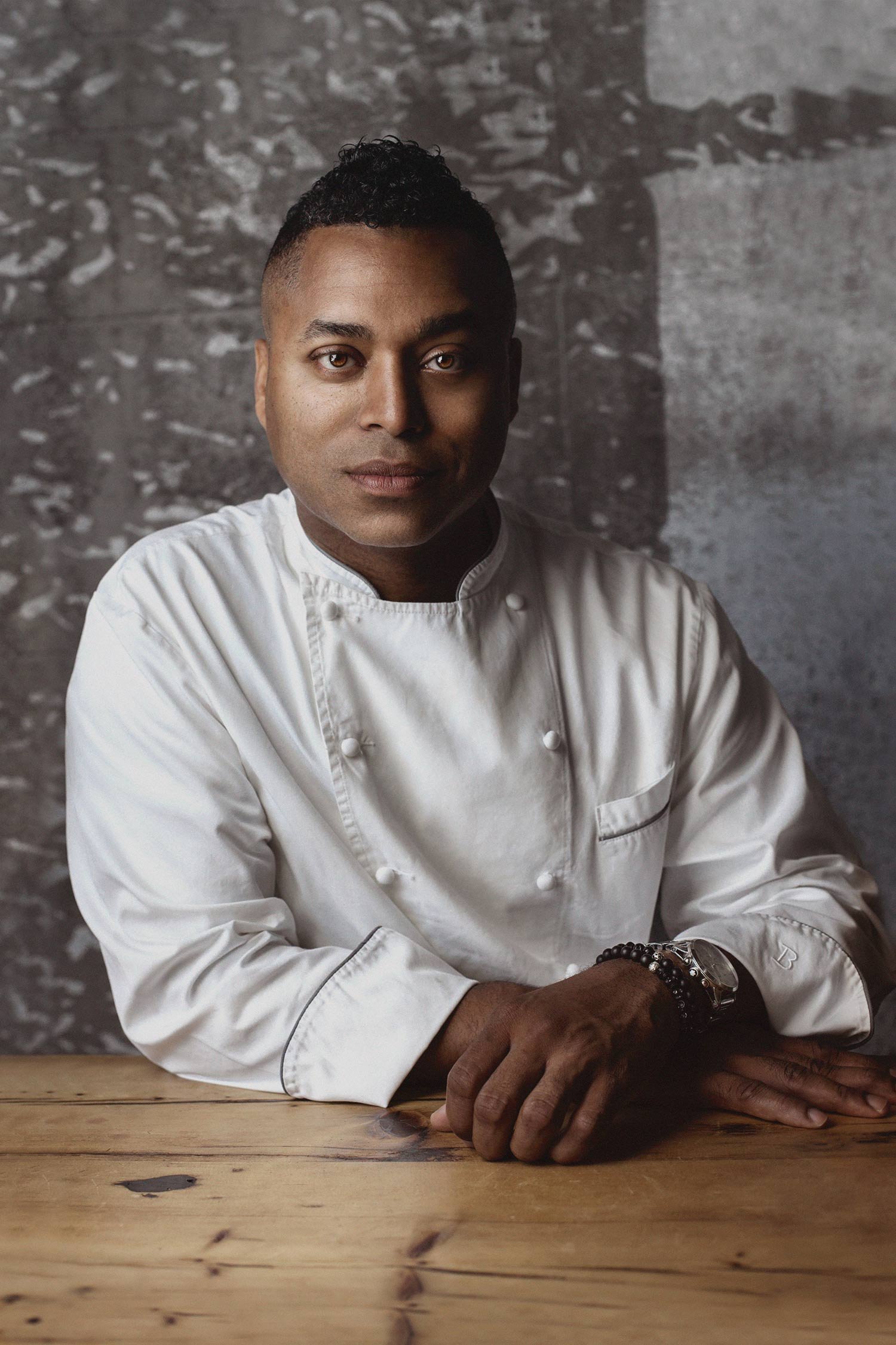 A male chef in a white chef's coat, sitting at a wooden table against a textured gray wall, looking at the camera.