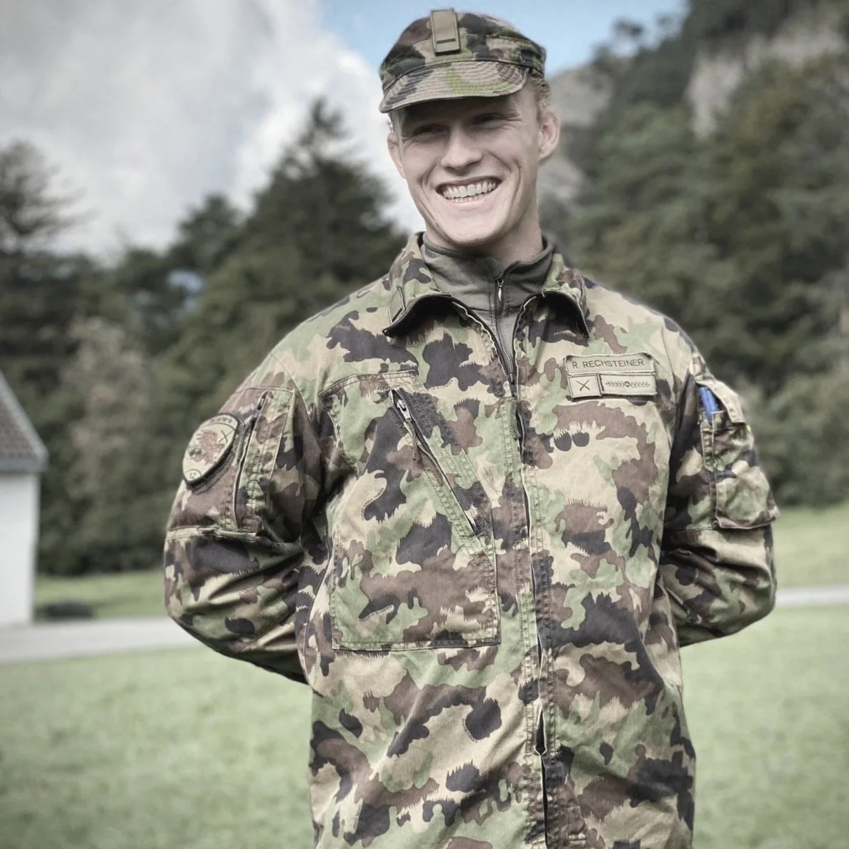 A smiling soldier wearing camouflage military uniform outdoors, with trees and a building in the background.