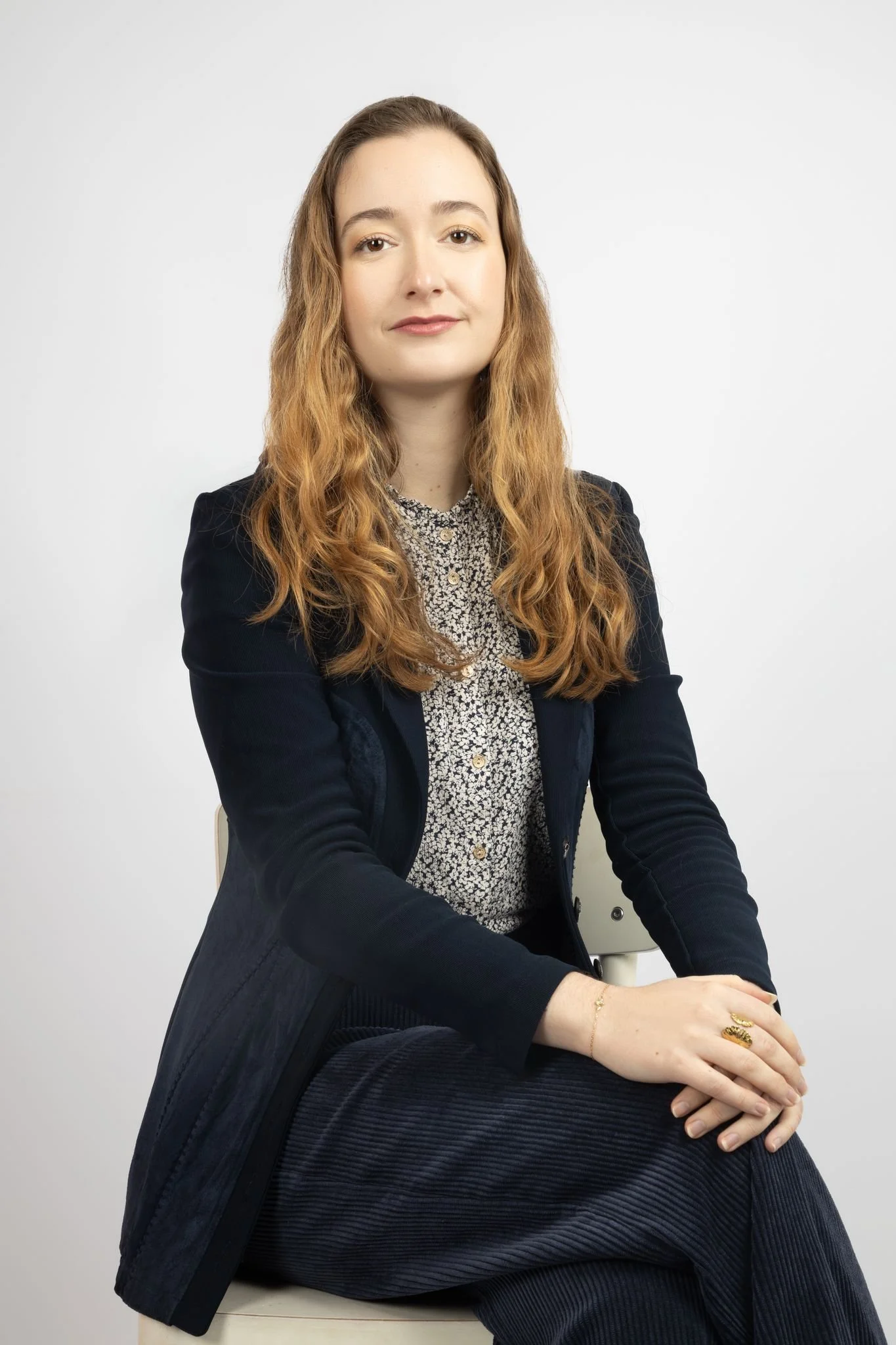 A young woman with long wavy red hair sitting on a chair against a plain white background, wearing a black blazer over a floral blouse and dark pants.