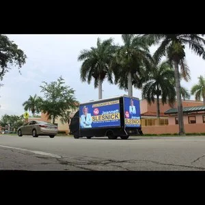 Mobile billboard truck carrying political campaign advertising on a city street lined with palm trees