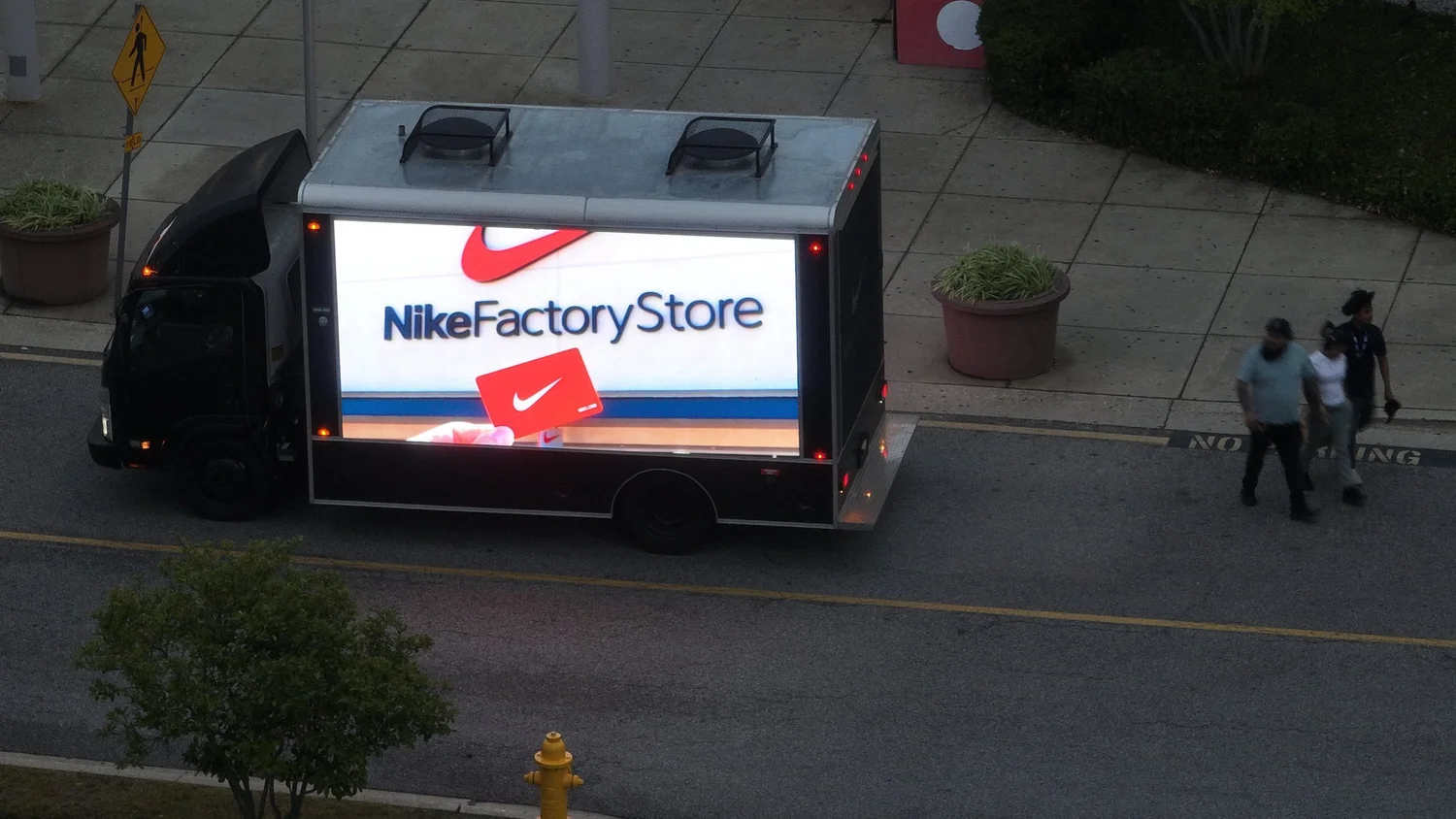 3-sided digital LED billboard truck displaying a Nike Factory Store ad on its side screen beside a city sidewalk