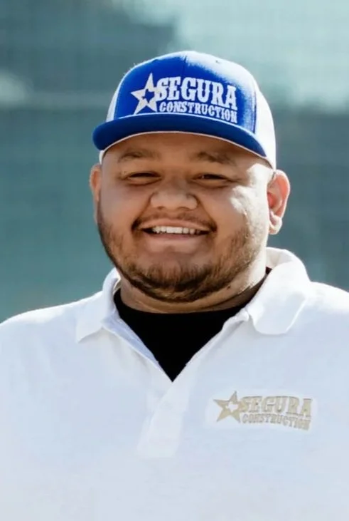 Smiling man wearing a blue cap with 'Segura Construction' logo and a white polo shirt with the same logo.