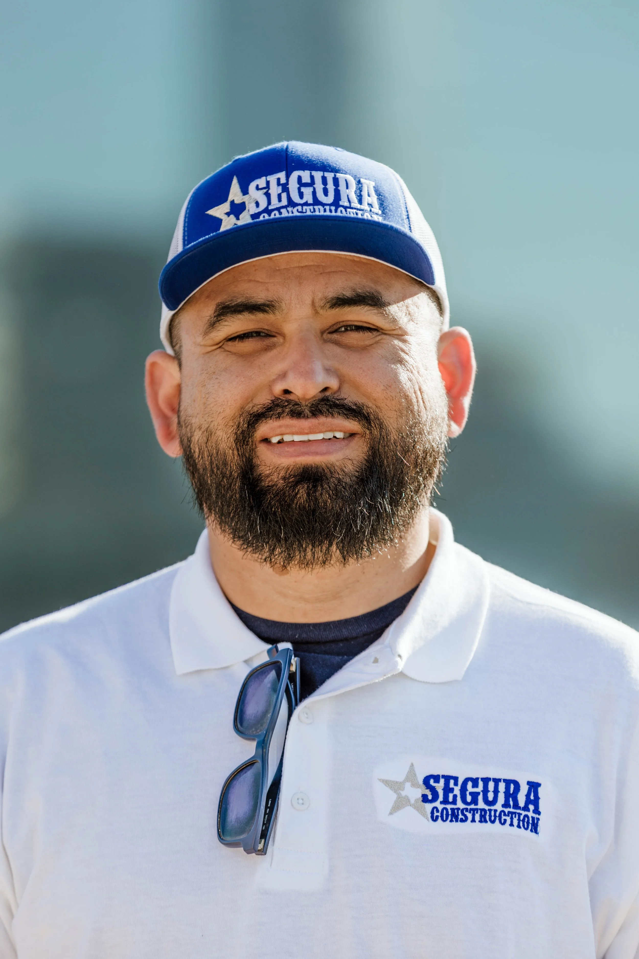 A man wearing a white polo shirt with Segura Construction logo, sunglasses hanging from the shirt collar, and a blue and white cap with the same logo, outdoors on a sunny day.