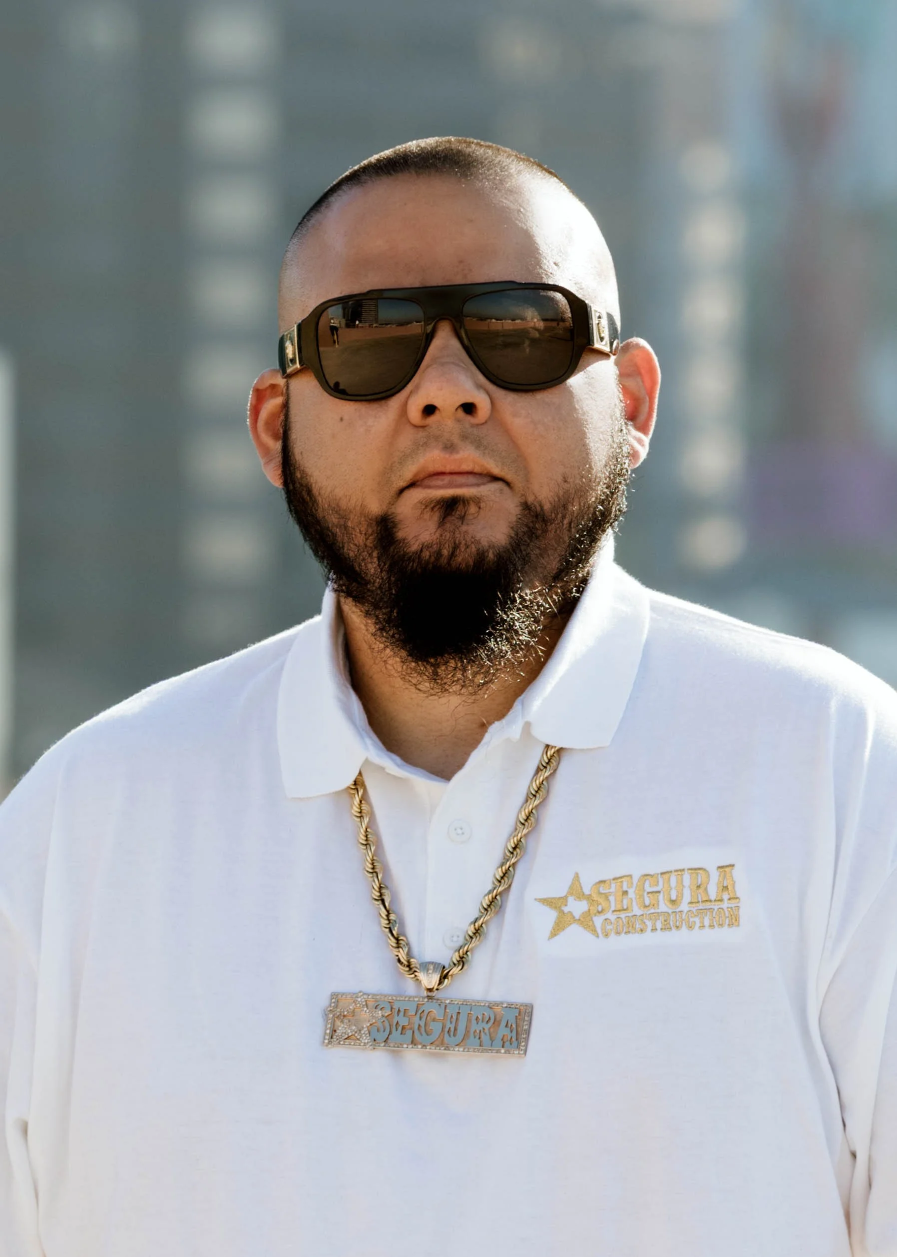 Man wearing black sunglasses, white polo shirt with 'Segura Construction' logo, gold chain, and 'Segura' nameplate necklace, standing outdoors in front of a cityscape.