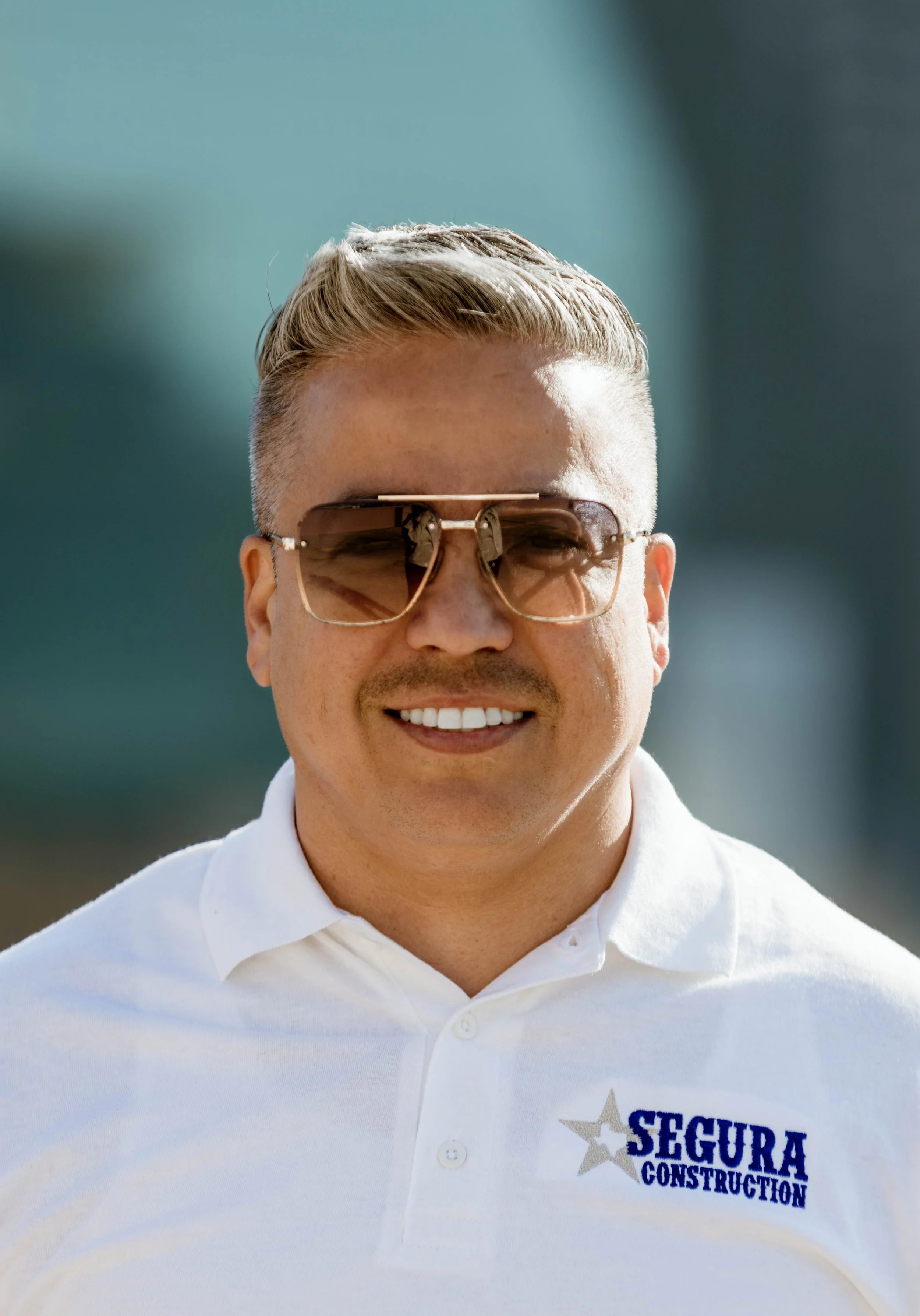 A man wearing sunglasses and a white polo shirt with the logo 'Segura Construction' smiling outdoors