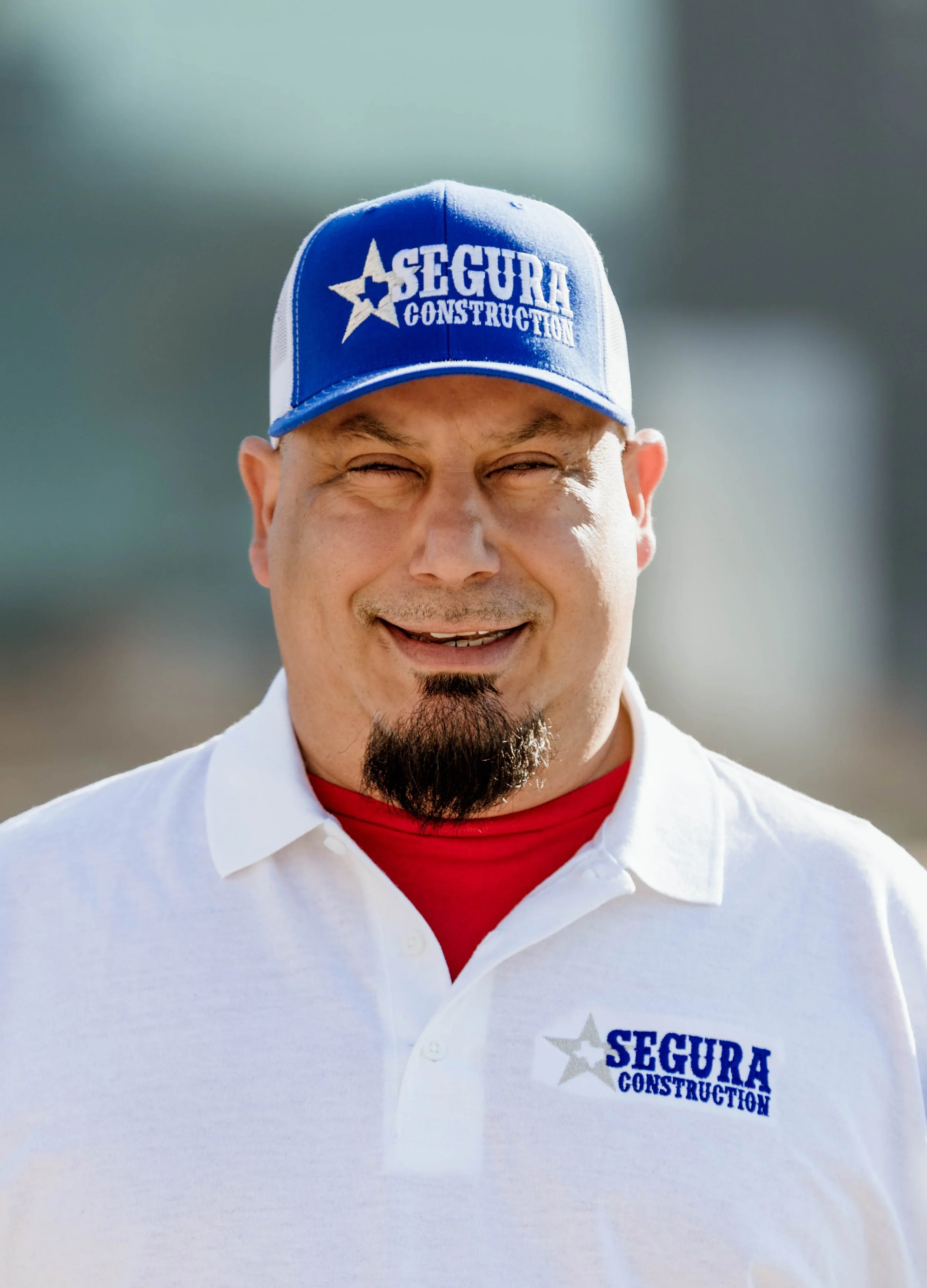 Man in white polo shirt and blue cap smiling outdoors, with 'Segura Construction' logo.