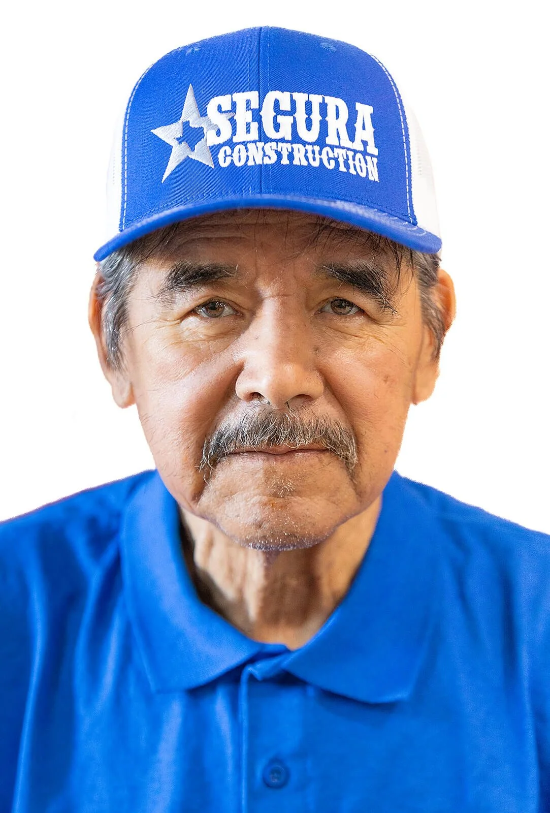 Close-up of an older man with a mustache wearing a blue cap with the logo 'Segura Construction' and a blue collared shirt.