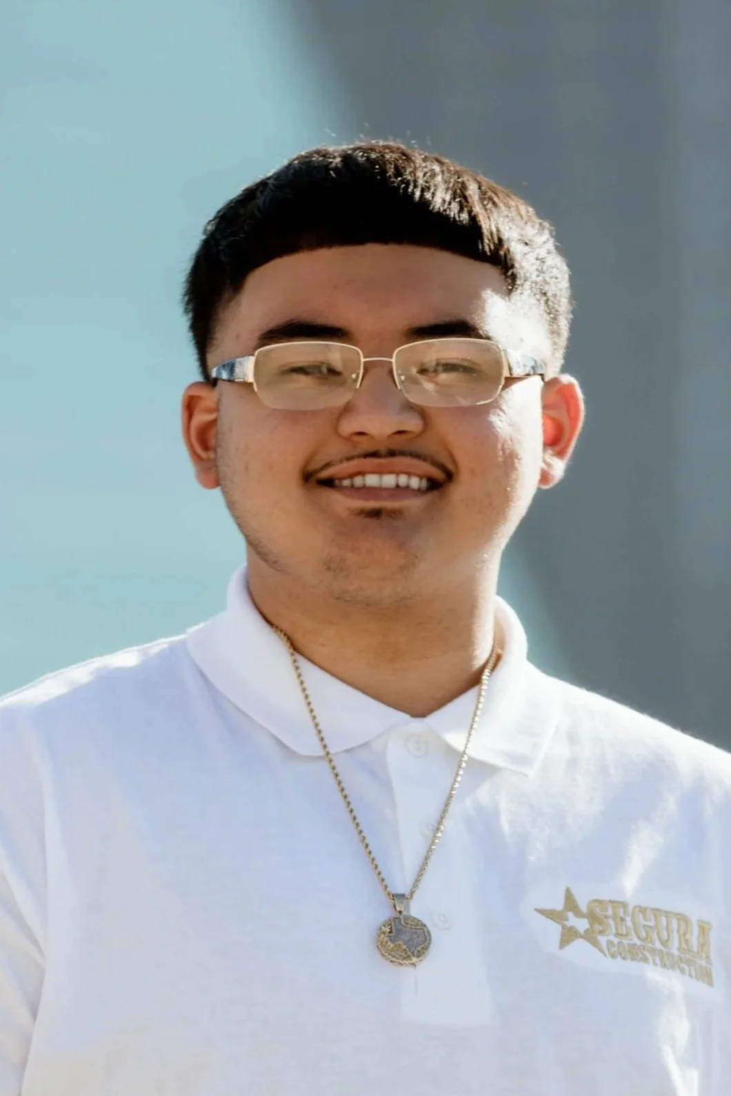 A young man with short black hair, glasses, and a goatee, smiling, wearing a white polo shirt with a 'Security Construction' logo and a gold chain necklace with a pendant.