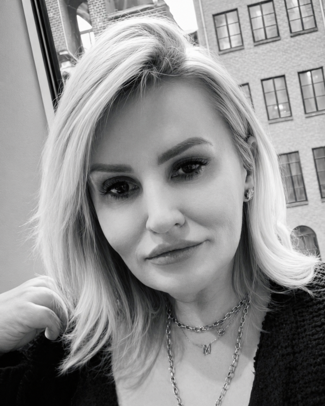 A black and white close-up photo of a woman with shoulder-length blonde hair, wearing layered necklaces and earrings, sitting near a window with a brick building with multiple windows visible outside.