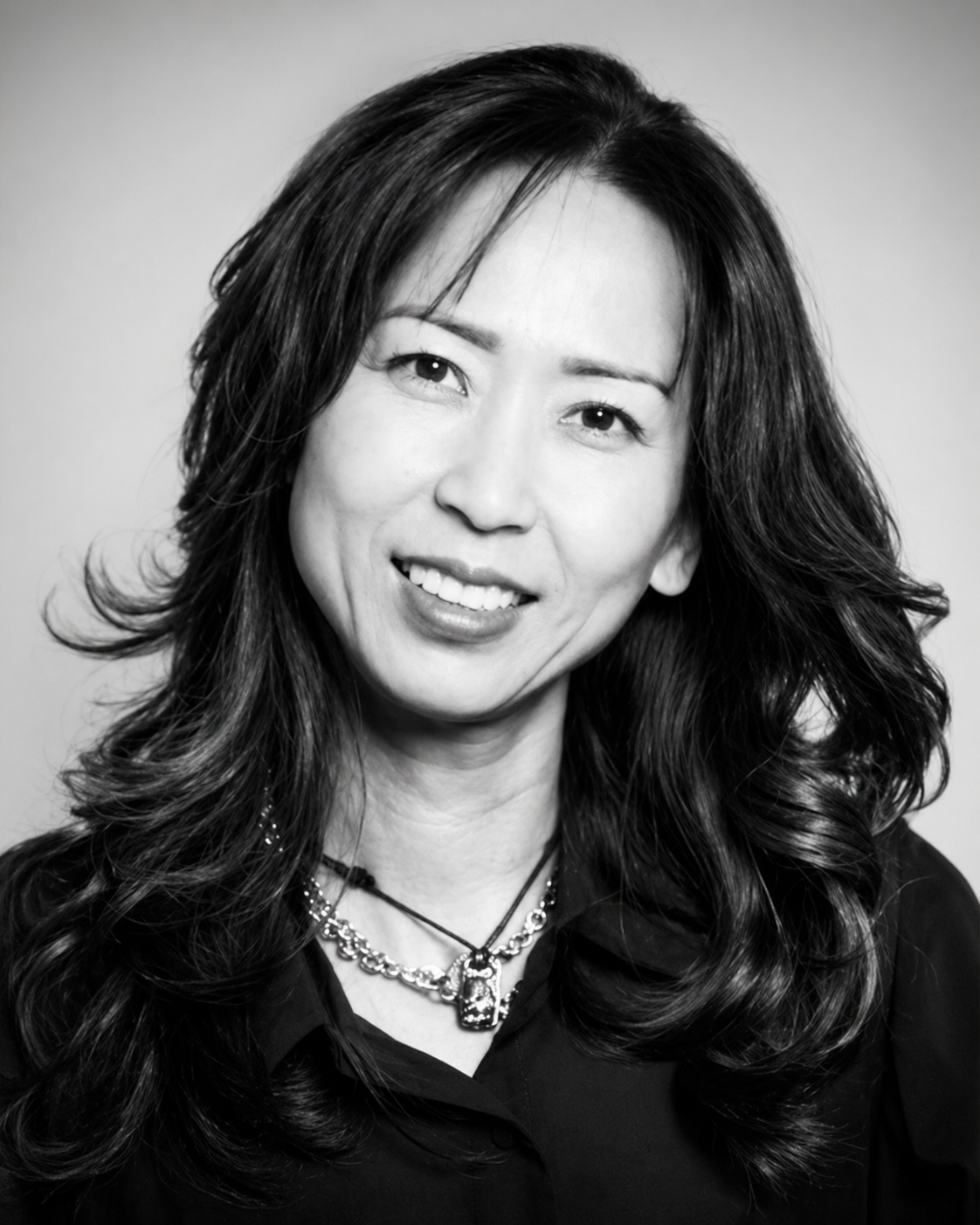 Black and white portrait of a woman with long wavy dark hair, smiling and wearing layered necklaces and a dark top.