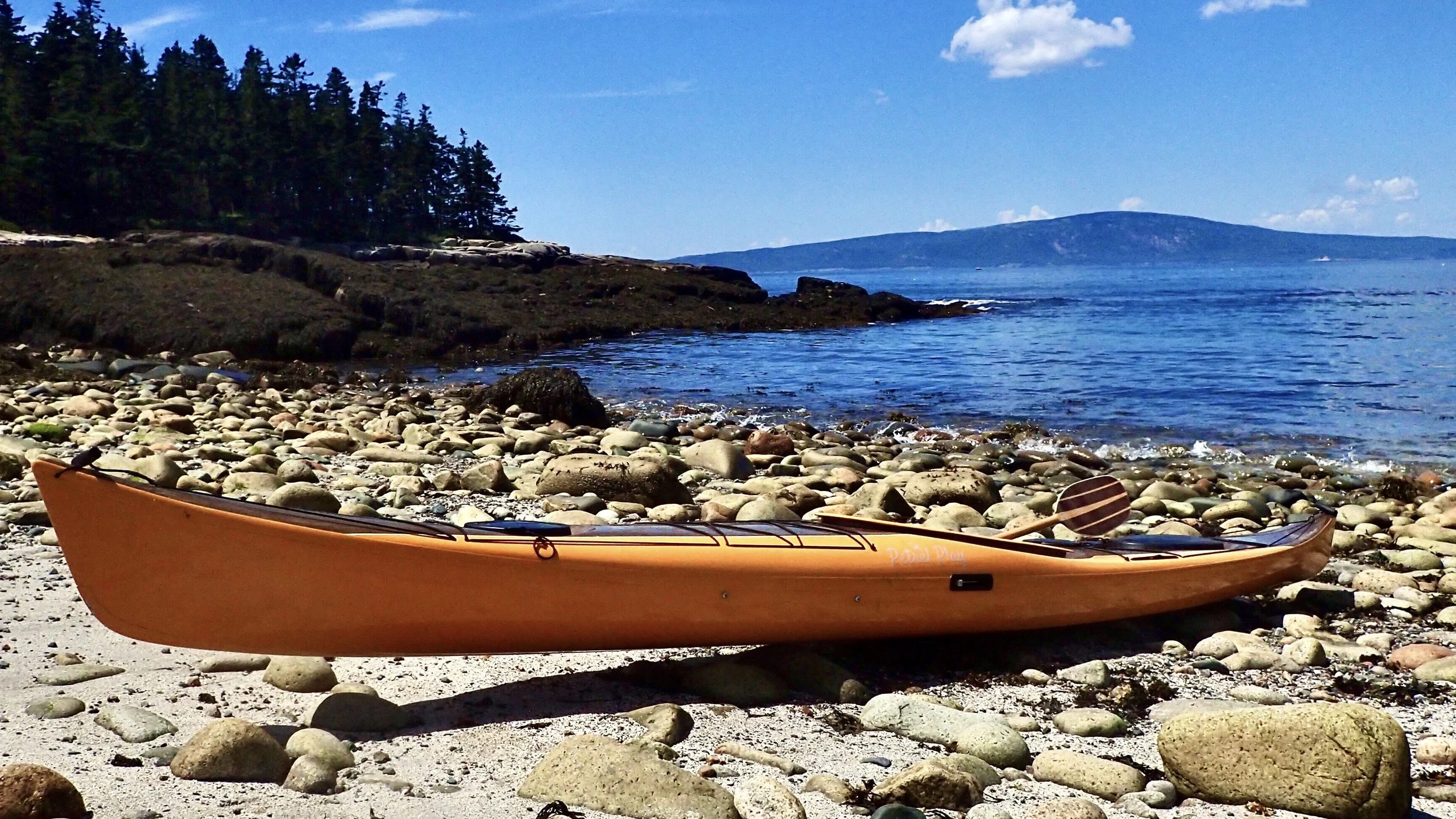 Orange composite kayak on rocky beach against ocean and forested hill backdrop