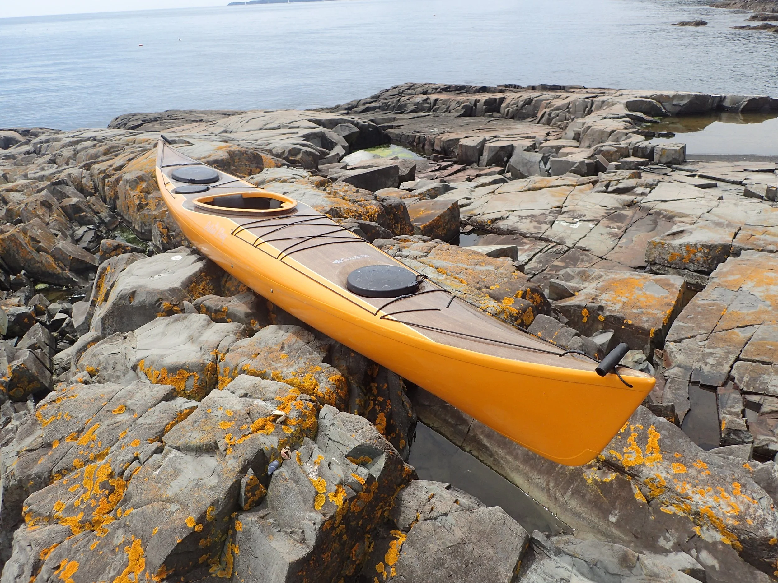 Yellow fiberglass and flax kayak resting on rocky shoreline with ocean in background.