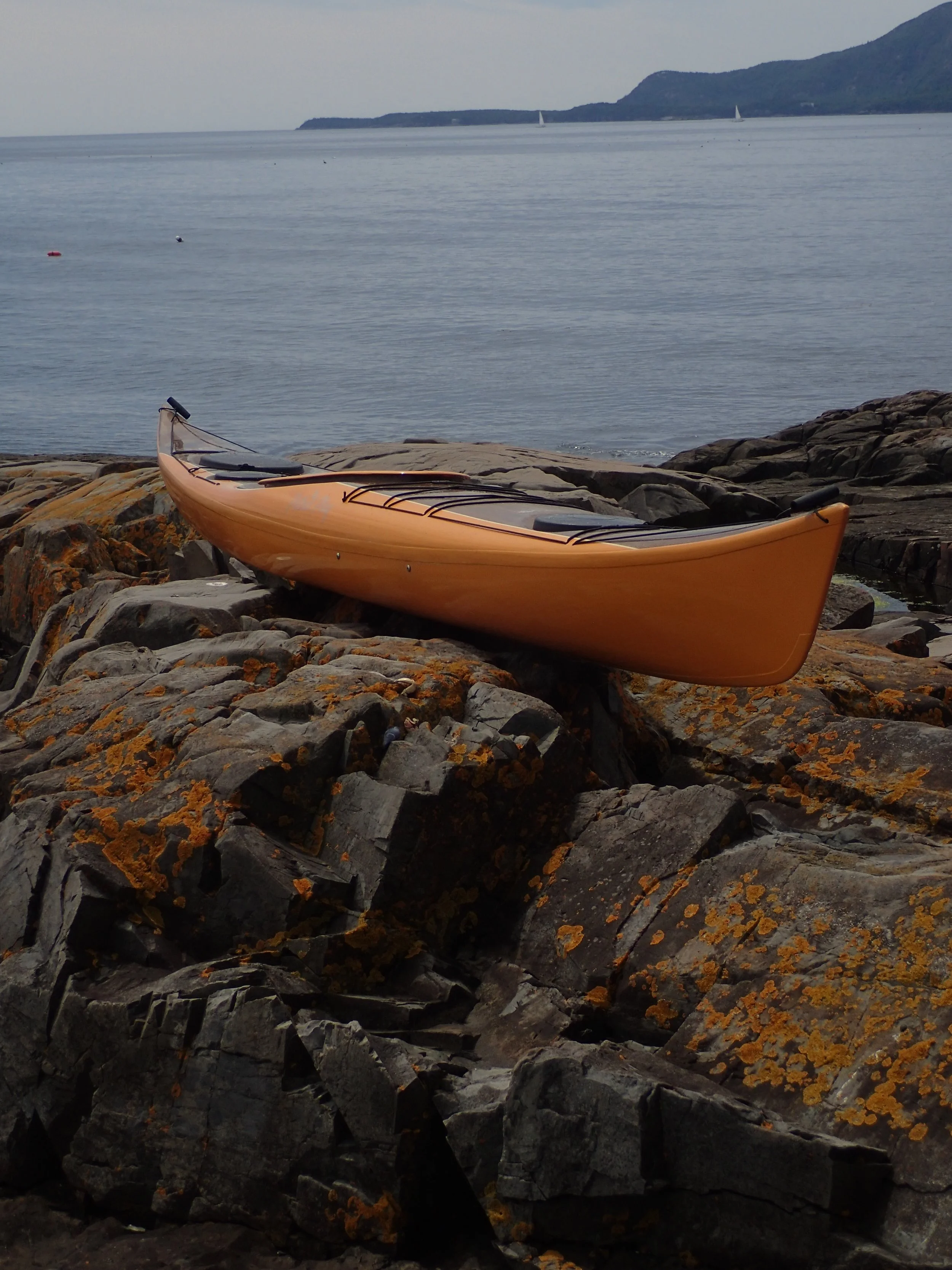 Orange infused composite kayak resting on rocky shore with lichen near calm sea and distant hills.