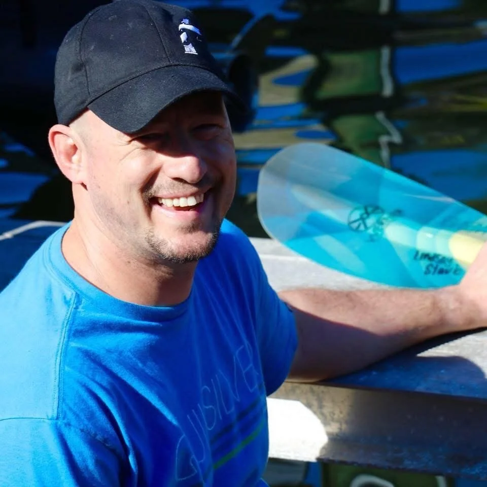 A smiling man wearing a black cap and blue shirt, sitting outdoors near a dock with rows of boats in the background, holding a clear boat windshield.