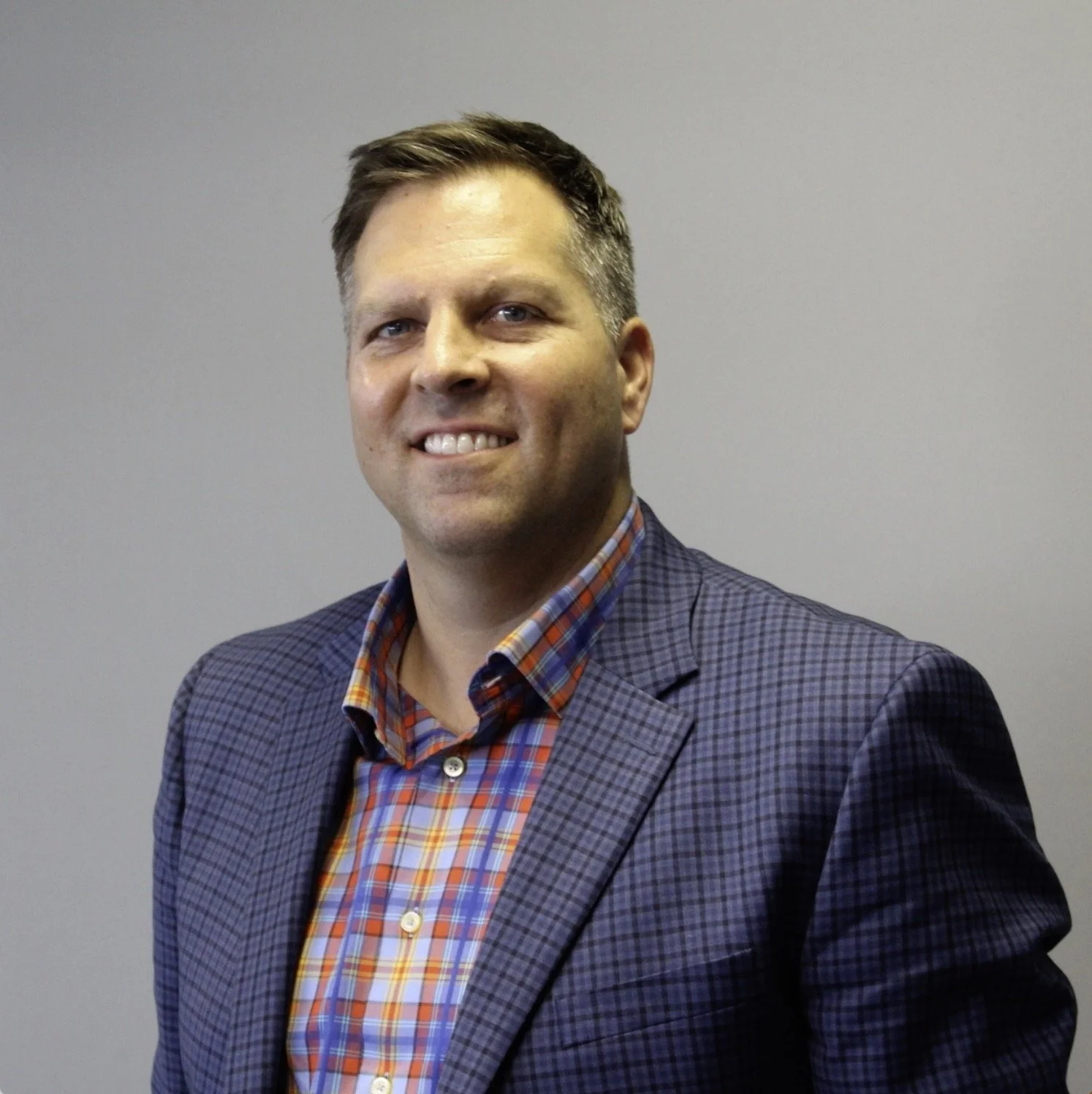 Man in a plaid shirt and blazer smiling, standing against a plain gray background.