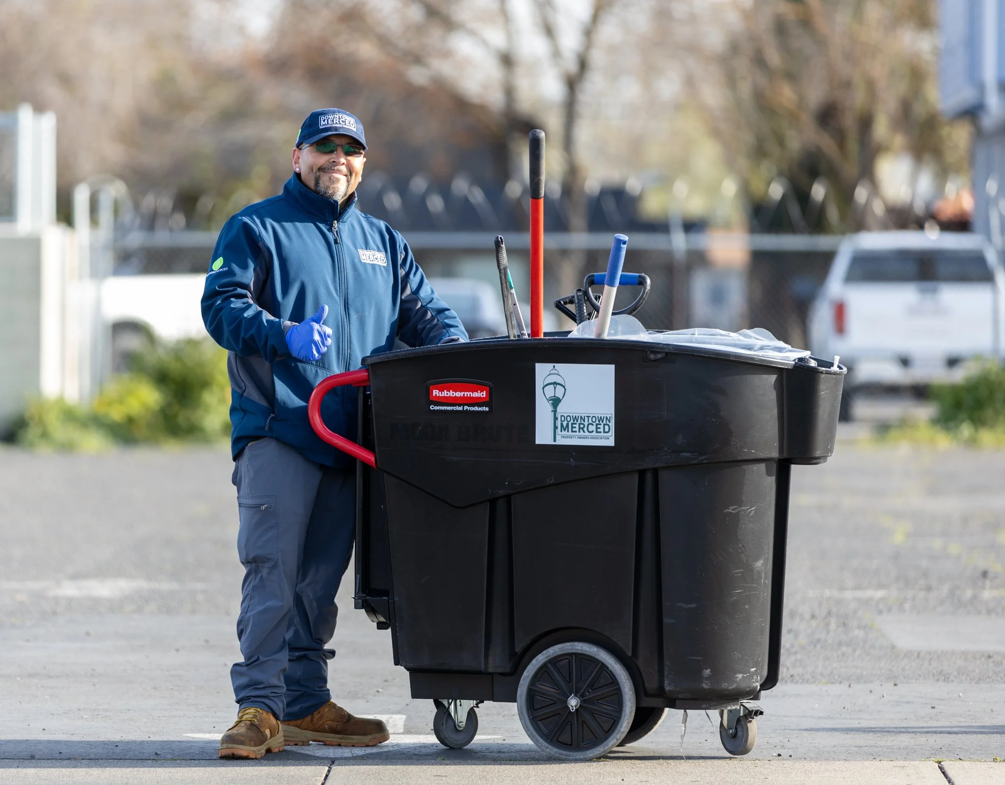 A man in a blue jacket and cap standing next to a black trash bin on wheels, smiling and giving a thumbs up. The bin has a sign that says 'Downtown Merced' and 'Rubbermaid'. The background shows a parking lot and trees.