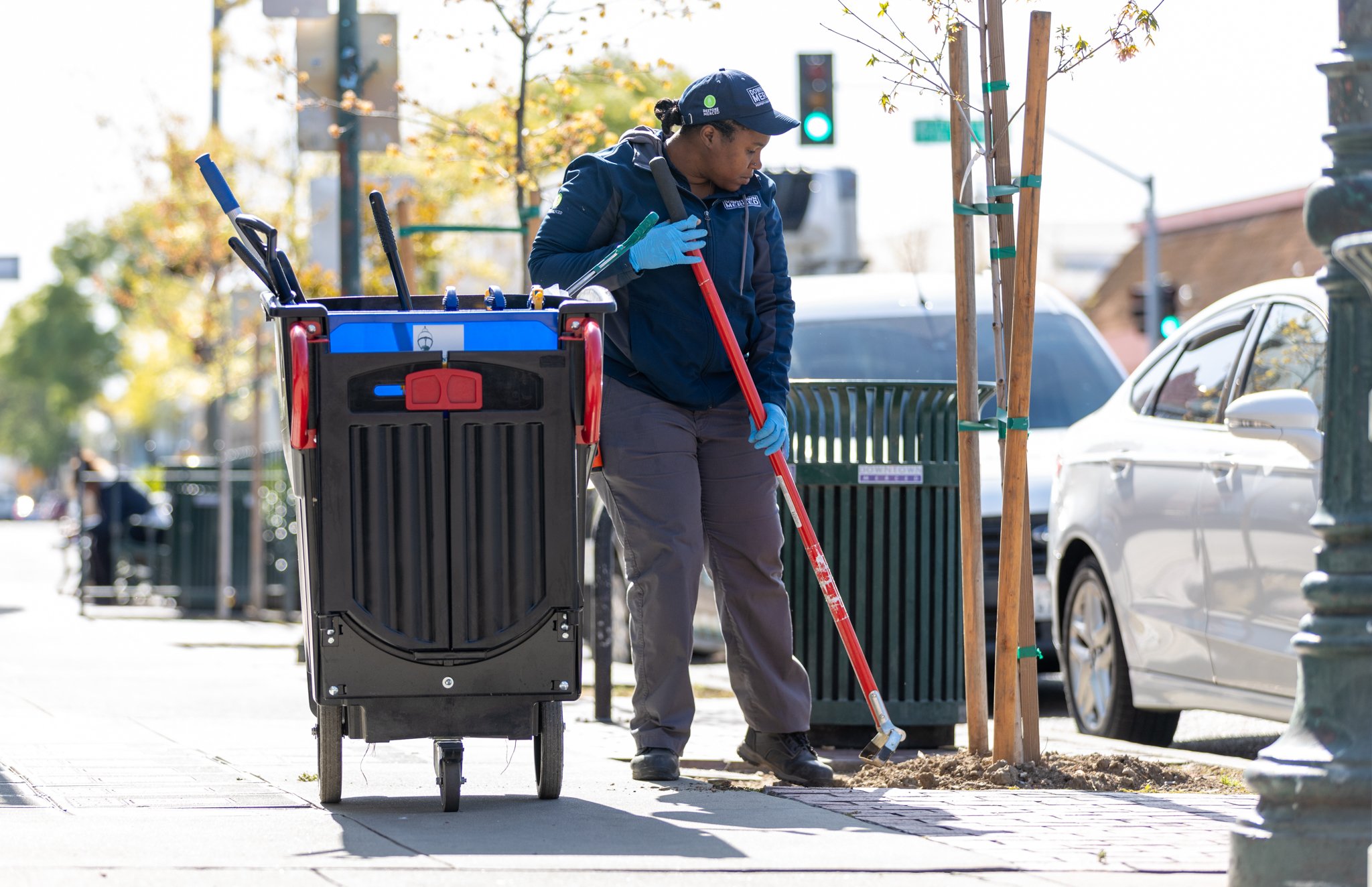 A worker sweeping the sidewalk next to a young tree with stakes in an urban area.