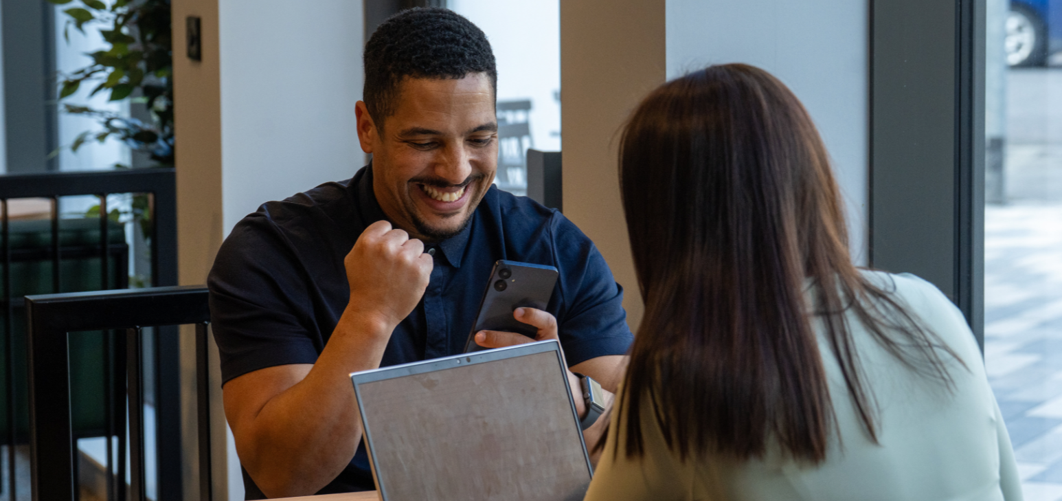 SG Global Expert Speakers - A man and woman sitting at a table in a cafe, smiling and looking at a smartphone.