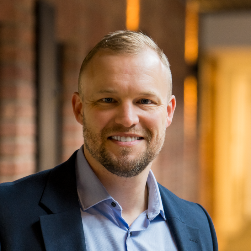 A smiling man with short blonde hair and a beard, wearing a blue blazer and light blue shirt, standing indoors in front of a brick wall with warm lighting.