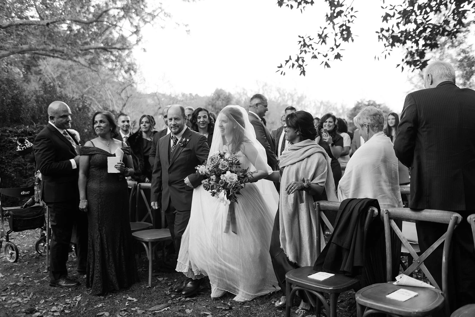 Bride walking down the aisle with a bouquet, surrounded by family and guests at an outdoor wedding ceremony.