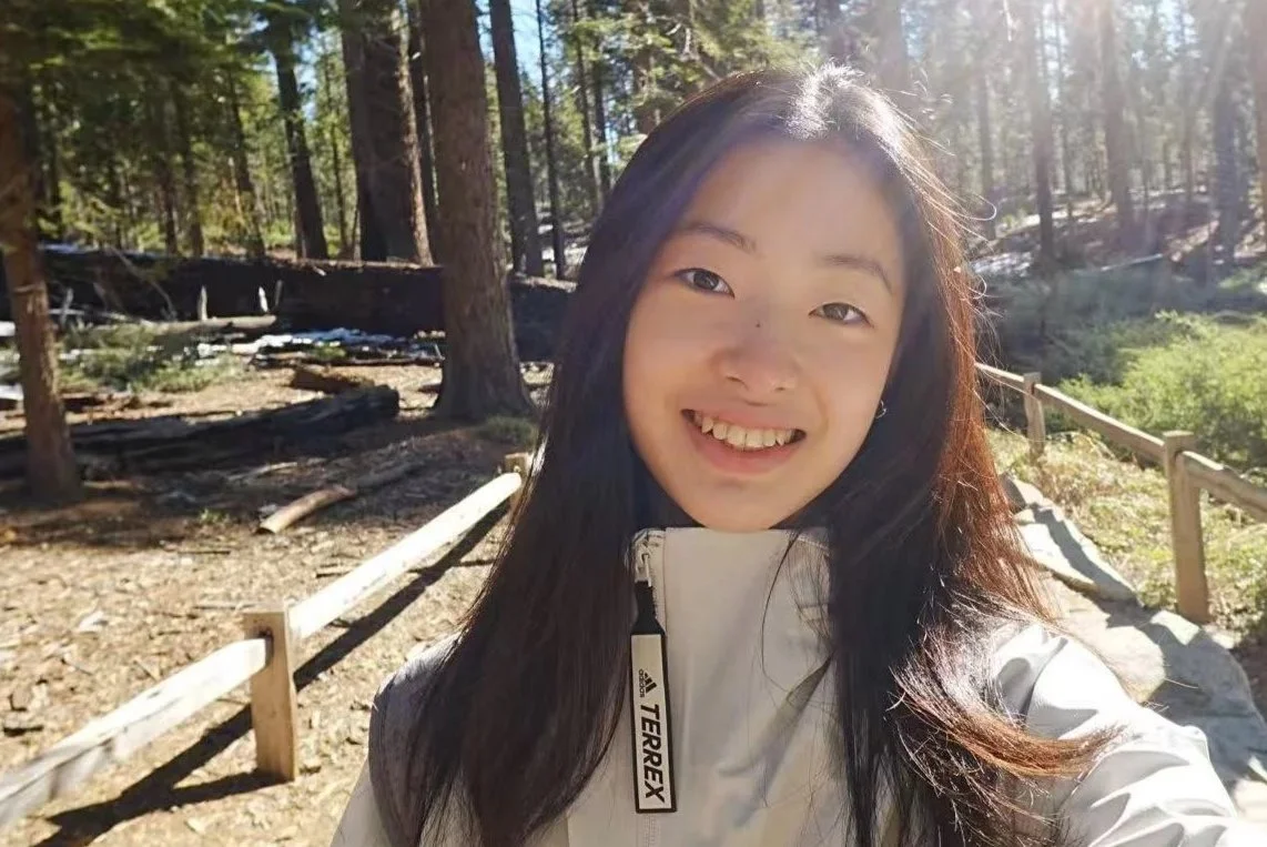 Smiling woman with long dark hair in a white jacket taking a selfie outdoors in a forest with trees and sunlight.